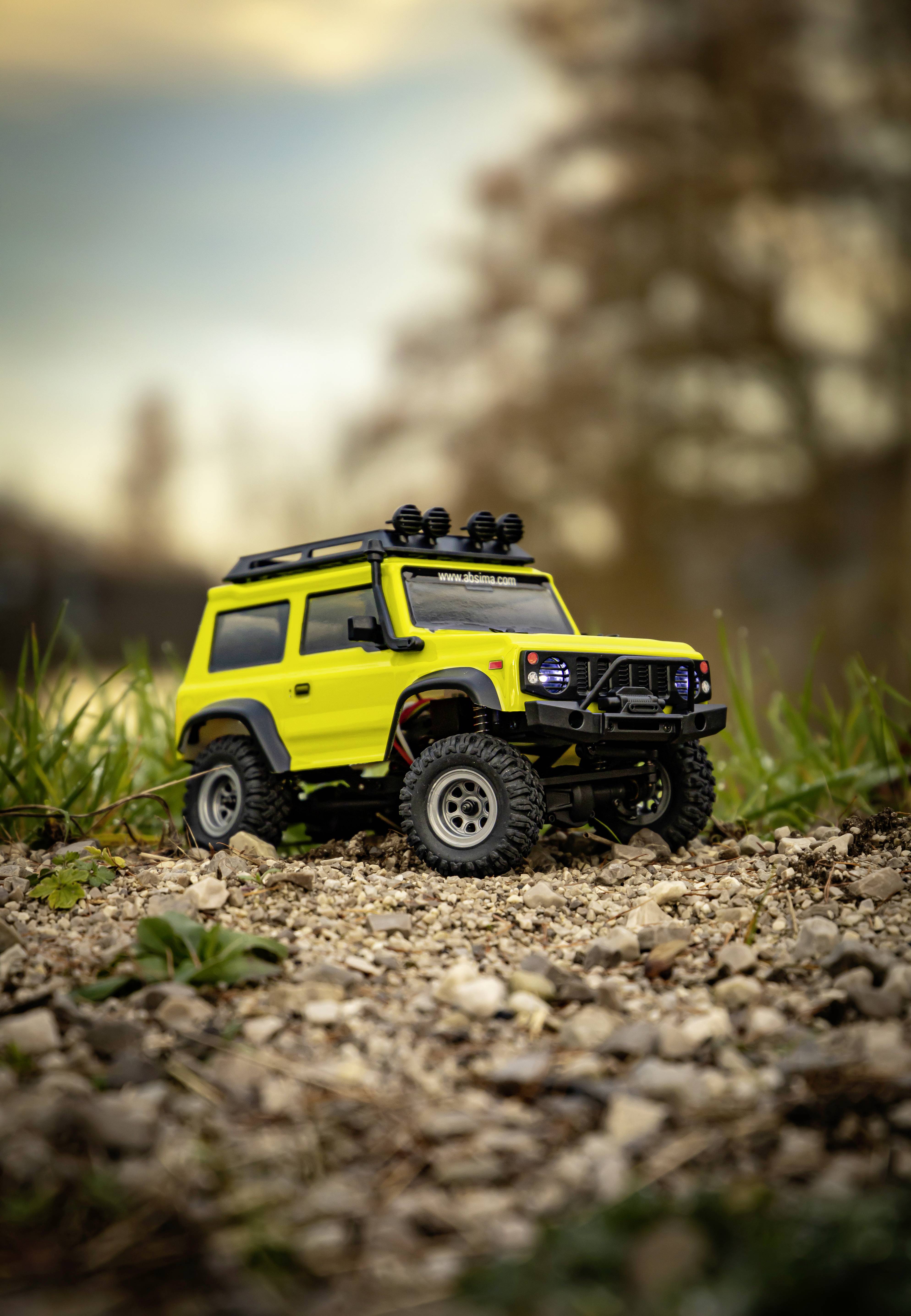 Ein leuchtend gelber Miniatur-Geländewagen steht auf einem kiesigen Weg, umgeben von Gras. Der Himmel ist bewölkt im Hintergrund.