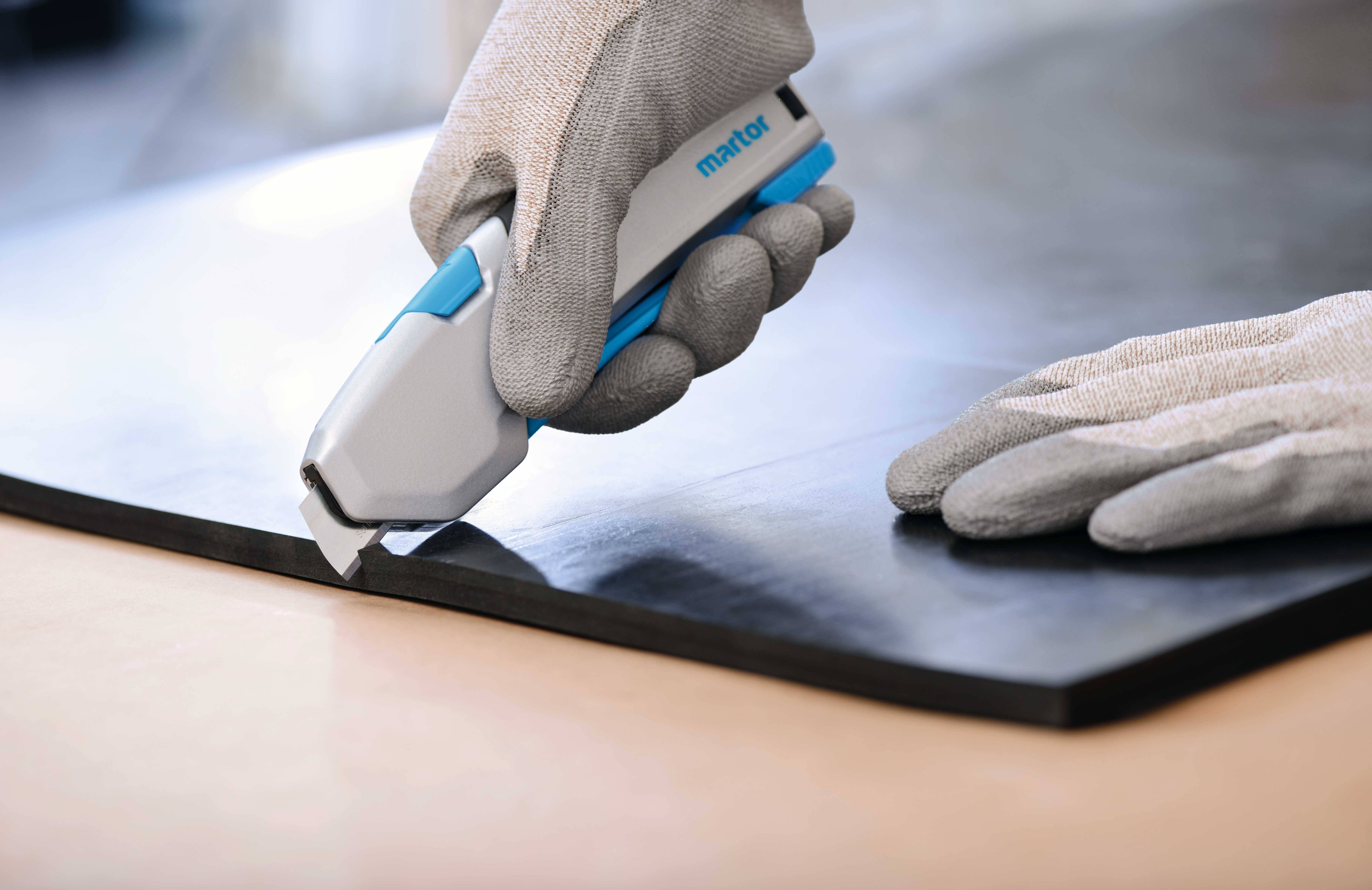 Ein Handschneider schneidet einen schwarzen Gummi auf einem Tisch. Ein Handschuh hält das Werkzeug für präzises Schneiden.