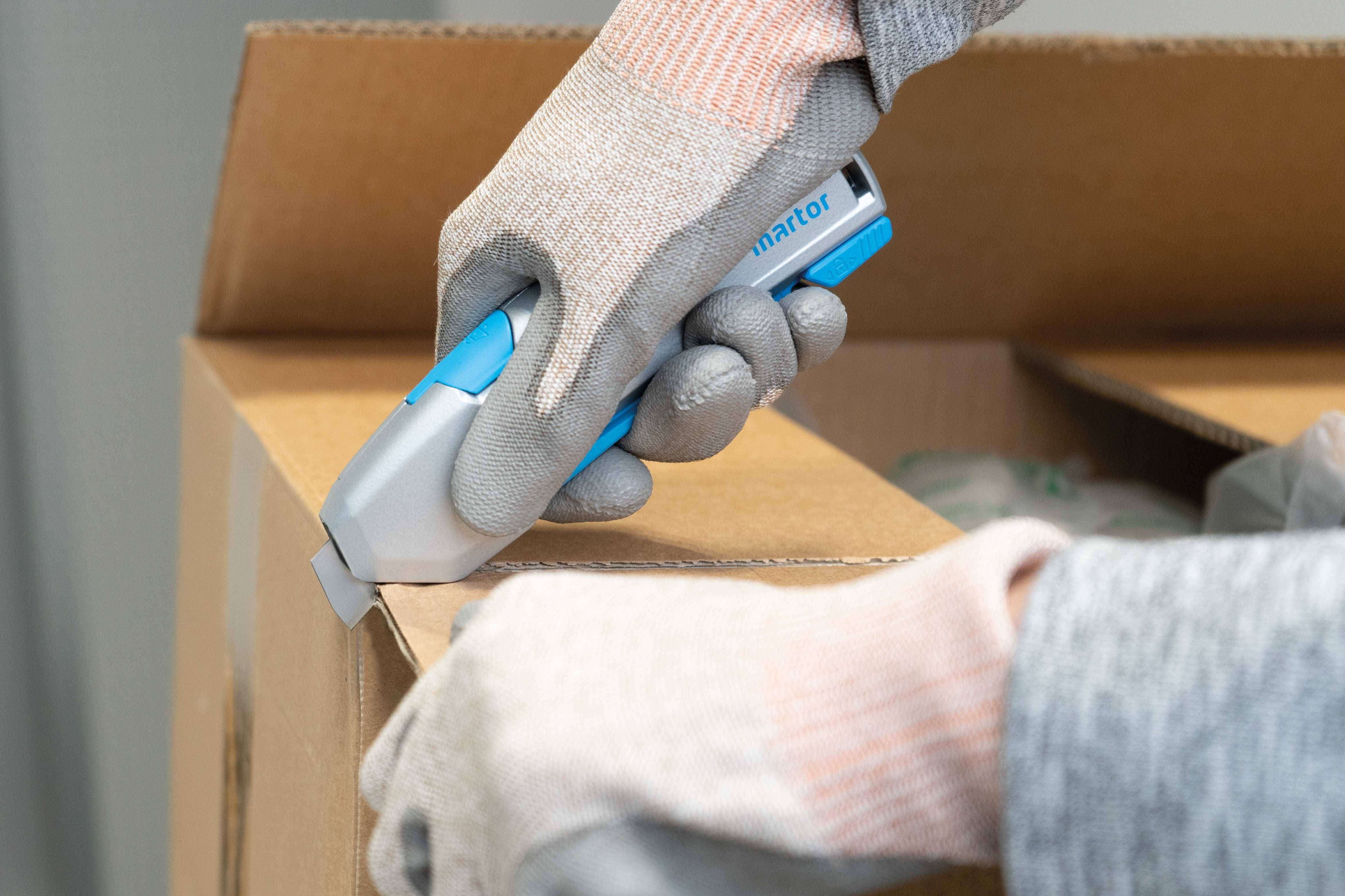 Eine Person mit grauen Sicherheits-Handschuhen öffnet einen Karton mit einem blauen Sicherheitsmesser.