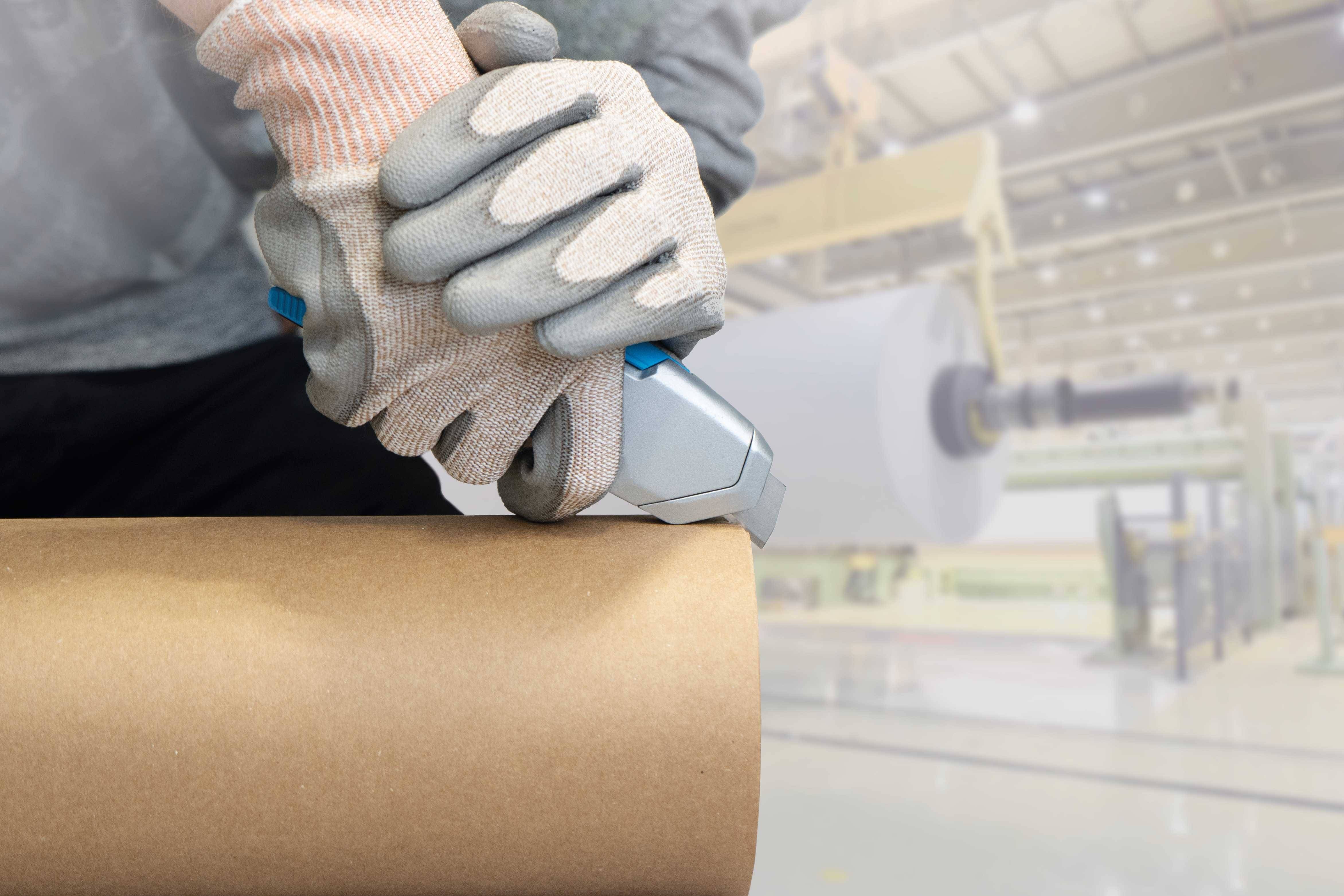 Eine Person trägt Handschuhe und schneidet eine Rolle Papier in einer Fabrikhalle. Im Hintergrund sind große Papierrollen sichtbar.