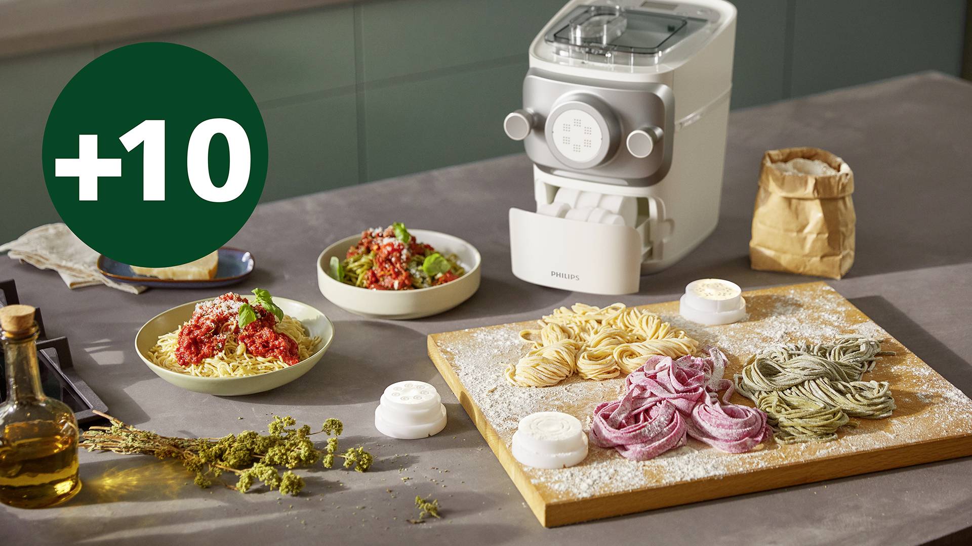 Frische, farbige Nudeln auf einem Brett, ein automatischer Pastamaker und Zutaten wie Olivenöl und Kräuter auf einem Küchentisch.