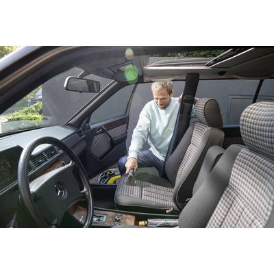 Ein Mann reinigt den Innenraum eines Autos, saugt den Boden. Er trägt einen hellblauen Pullover. Tageslicht fällt durch das Autofenster.