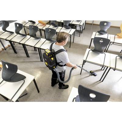Mann mit Rucksackstaubsauger reinigt einen leeren Klassenraum. Stühle sind hochgestellt, der Boden wird gesaugt.