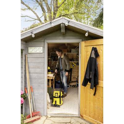 Ein Mann reinigt ein Gartenhaus mit einem gelben Staubsauger. Geöffnete Tür, Werkzeuge und Pflanzen sind sichtbar. Holzhaus im Garten.