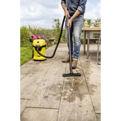 Eine Person benutzt einen gelben Nass-/Trockensauger auf einer Holzterrasse, um Wasser und Schmutz zu entfernen. Im Hintergrund sind ein Tisch und grüner Rasen zu sehen.