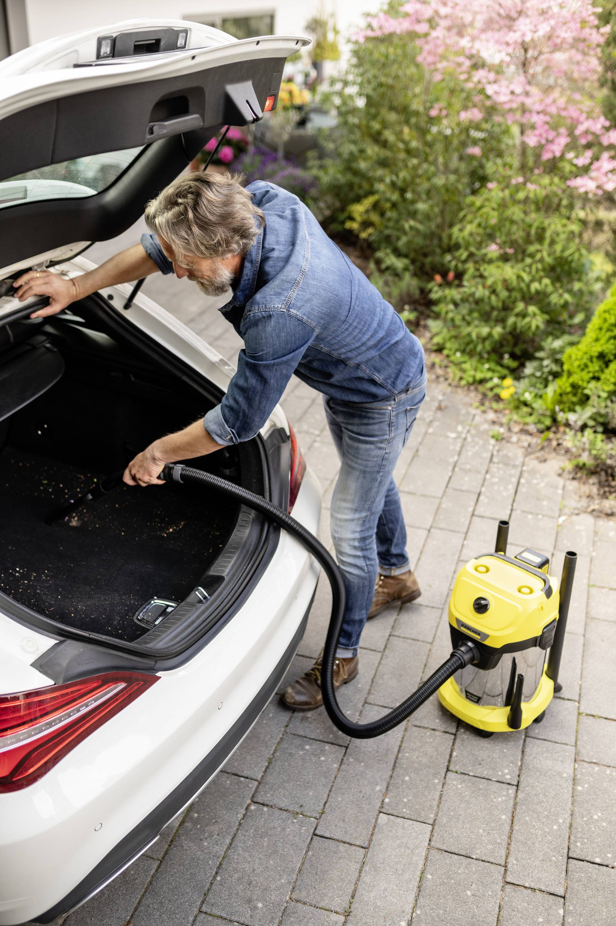 Ein Mann saugt den Kofferraum eines weißen Autos mit einem gelben Staubsauger aus, umgeben von Pflanzen im Freien.