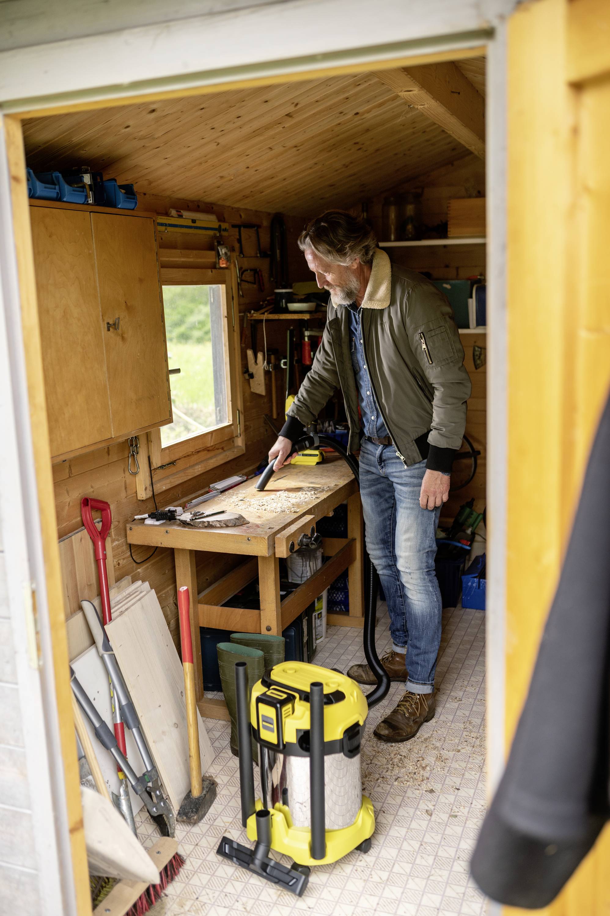 Ein Mann steht in einer Holzhütte und arbeitet an einer Werkbank. Werkzeuge und Geräte, darunter ein Staubsauger, sind um ihn herum zu sehen.