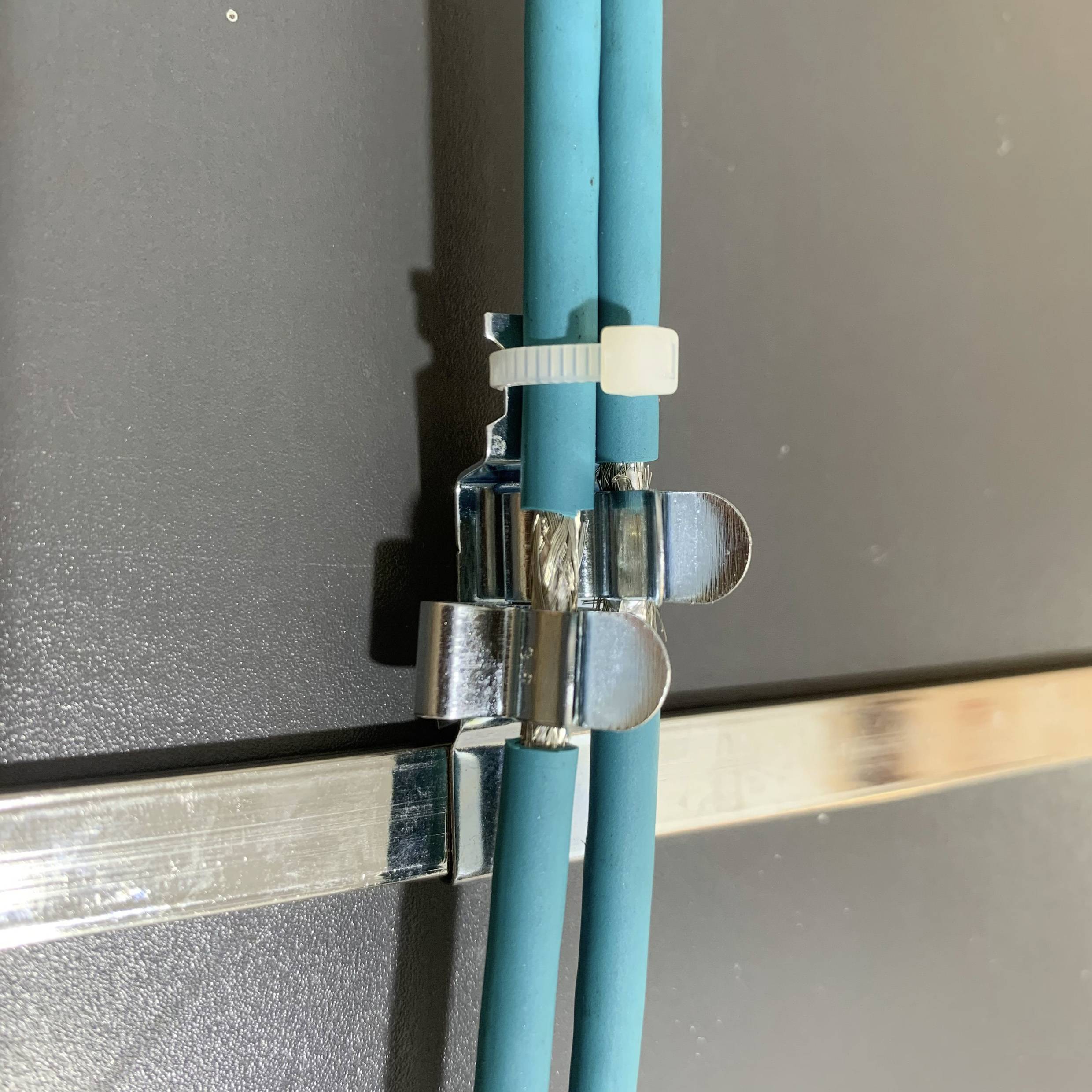 Ein Kabelmanagement-System zeigt zwei blaue Kabel, die an einer metallischen Oberfläche mit einer weißen Kabelschelle befestigt sind.