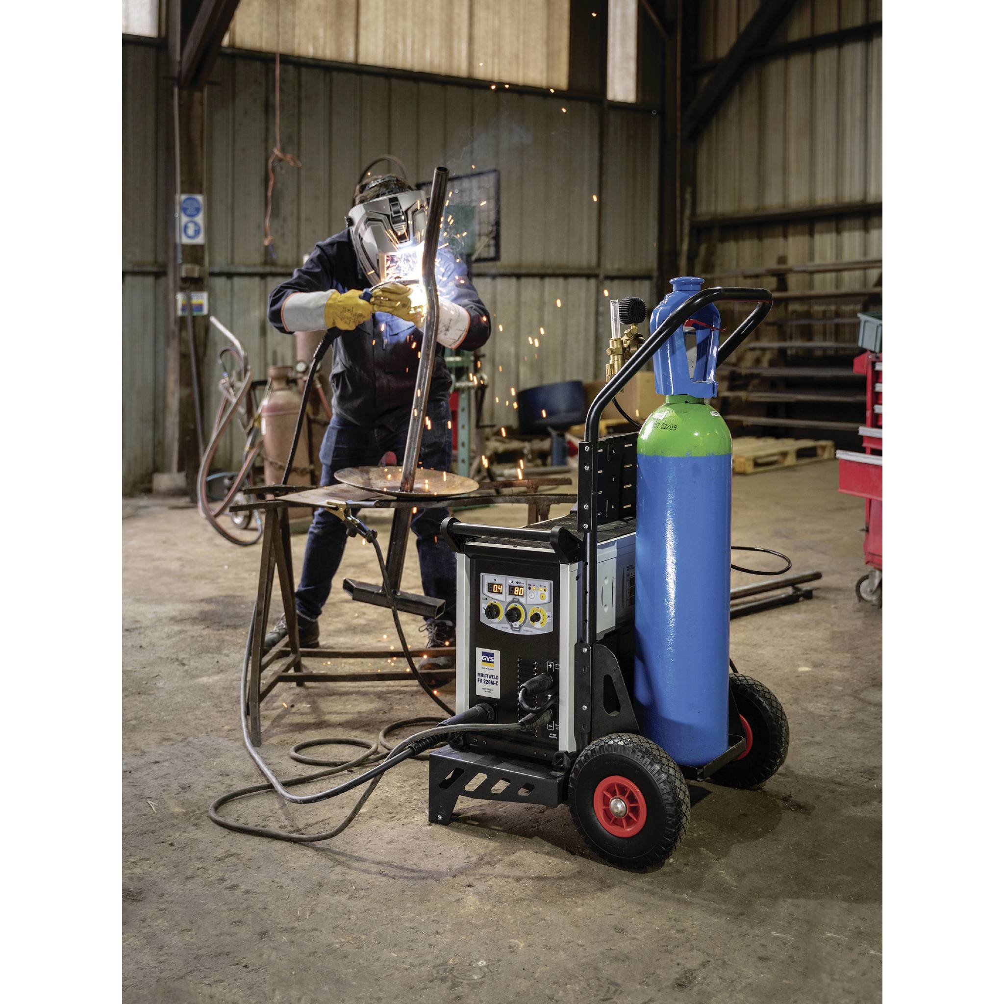 'Person beim Schweißen mit Schutzausrüstung in Werkstatt umgeben von Werkzeugen, Schweißgerät und Gasflasche im Vordergrund.'