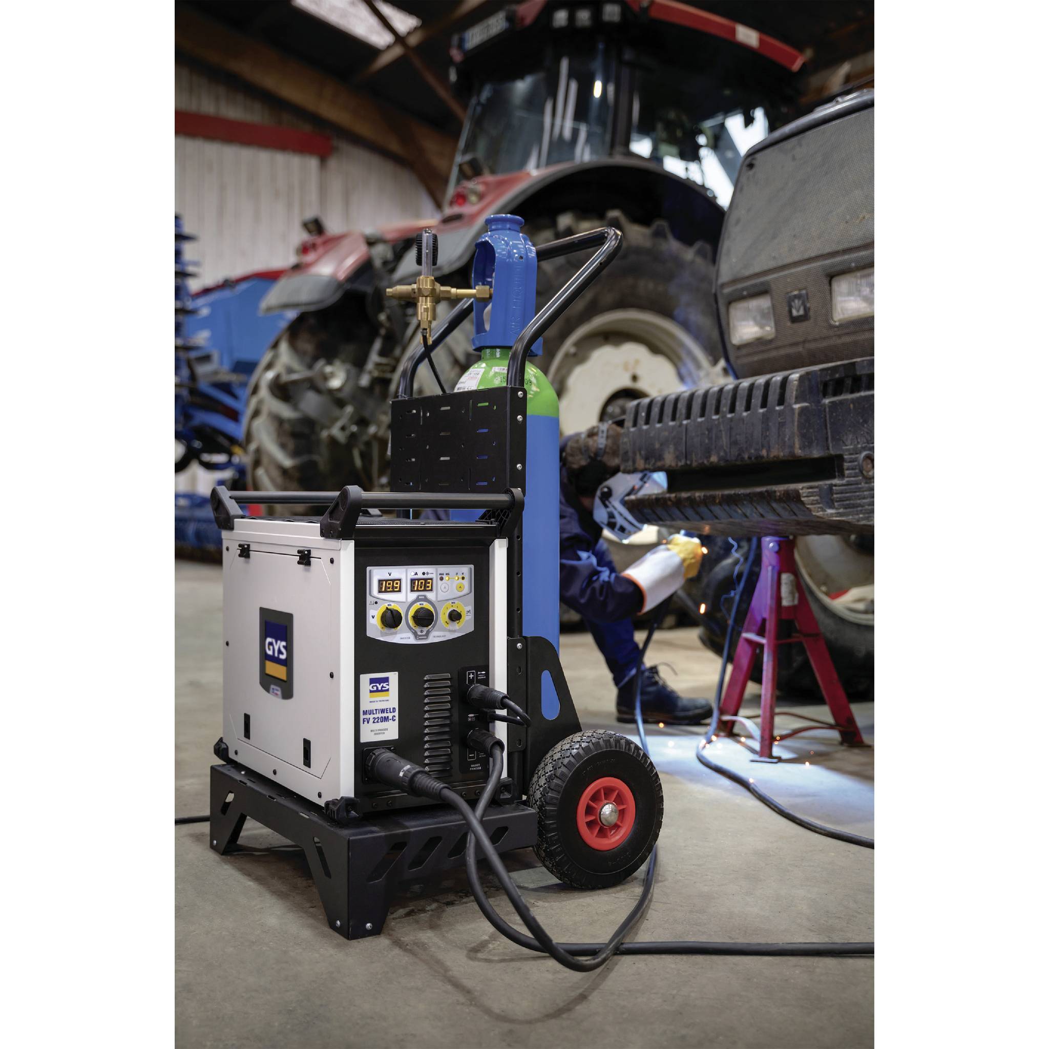 Ein Mechaniker repariert ein Fahrzeug, während ein Schweißgerät im Vordergrund steht. Im Hintergrund sind landwirtschaftliche Maschinen.