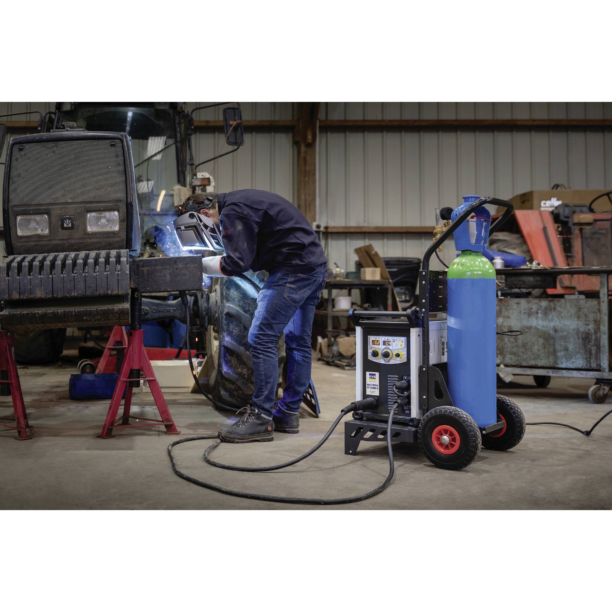 Eine Person schweißt in einer Werkstatt. Sie trägt Schutzkleidung und arbeitet mit einem Schweißgerät und Gasflasche in der Nähe eines großen Fahrzeugs.