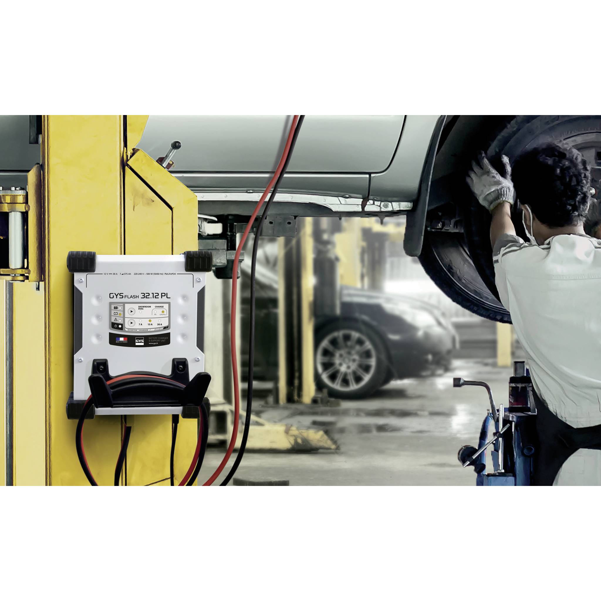 Ein Mechaniker arbeitet an einem angehobenen Auto in einer Werkstatt. Ein Batterieprüfgerät ist an einem Stützpfosten befestigt.