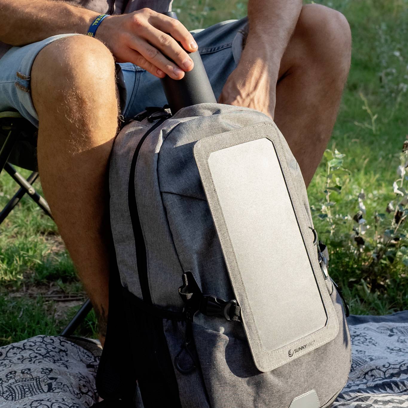 Eine Person sitzt im Freien auf einem Campingstuhl, hält eine Thermoskanne und hat einen grauen Rucksack mit Solarzellenpanel vorne.