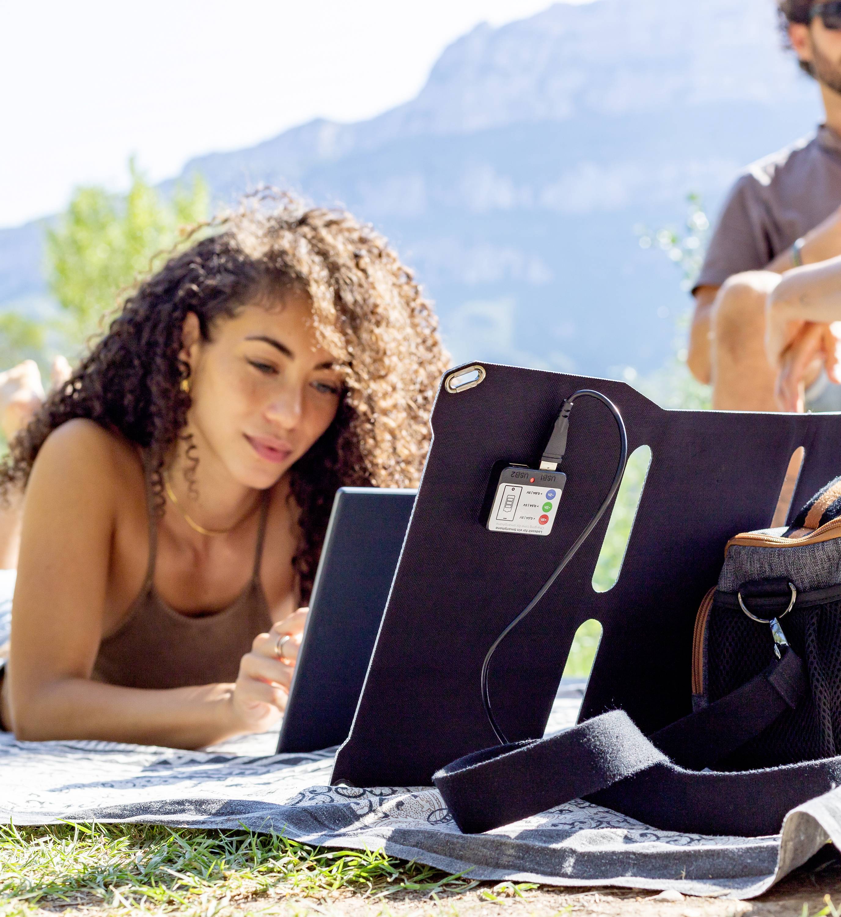 Eine Person liegt auf einer Decke im Freien und arbeitet an einem Laptop. Ein Berg ist im Hintergrund sichtbar.