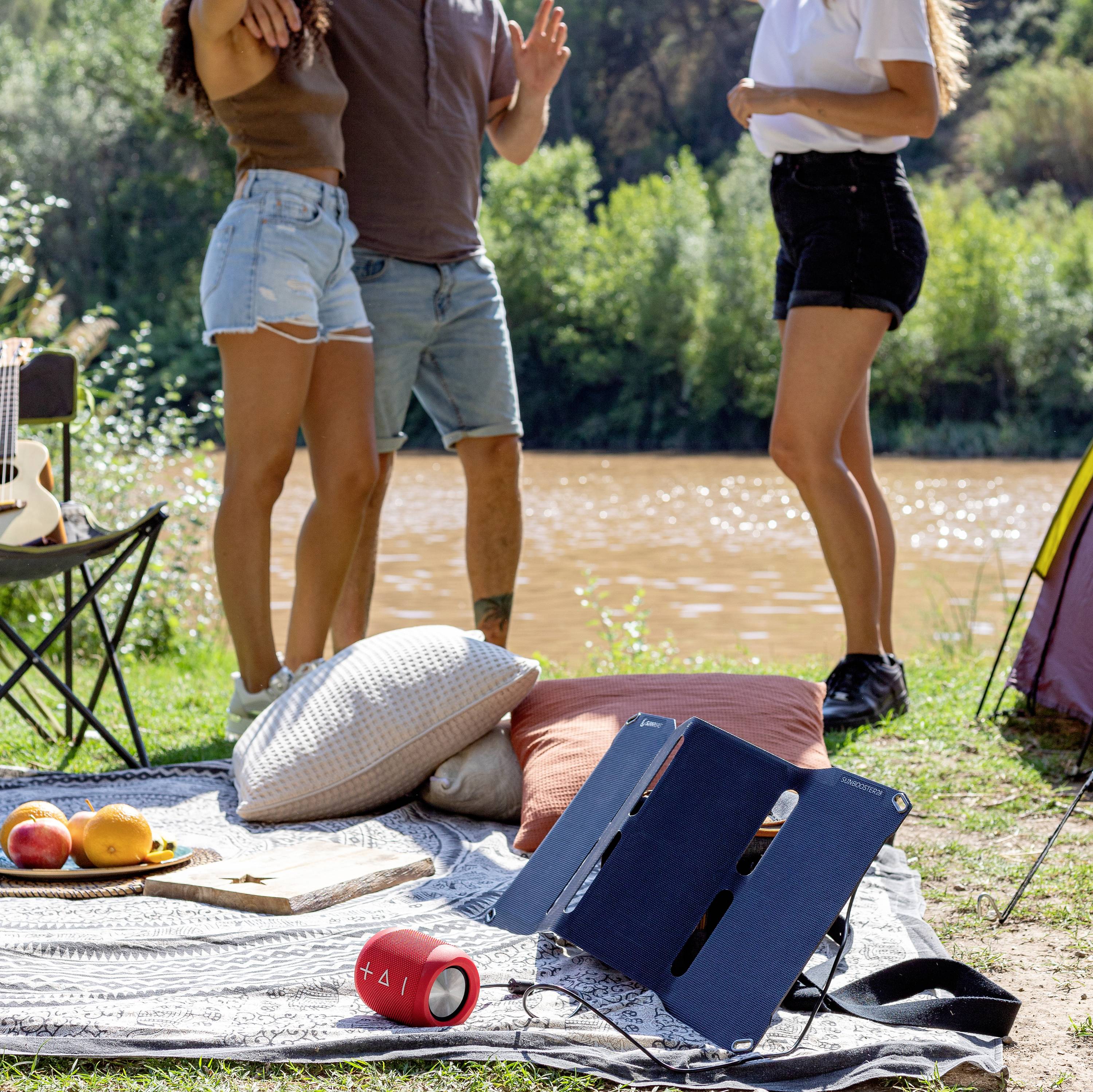 Drei Personen stehen lachend vor einem Fluss. Im Vordergrund ein Picknick mit Decke, Kissen, Obst, Bluetooth-Lautsprecher und Laptop.