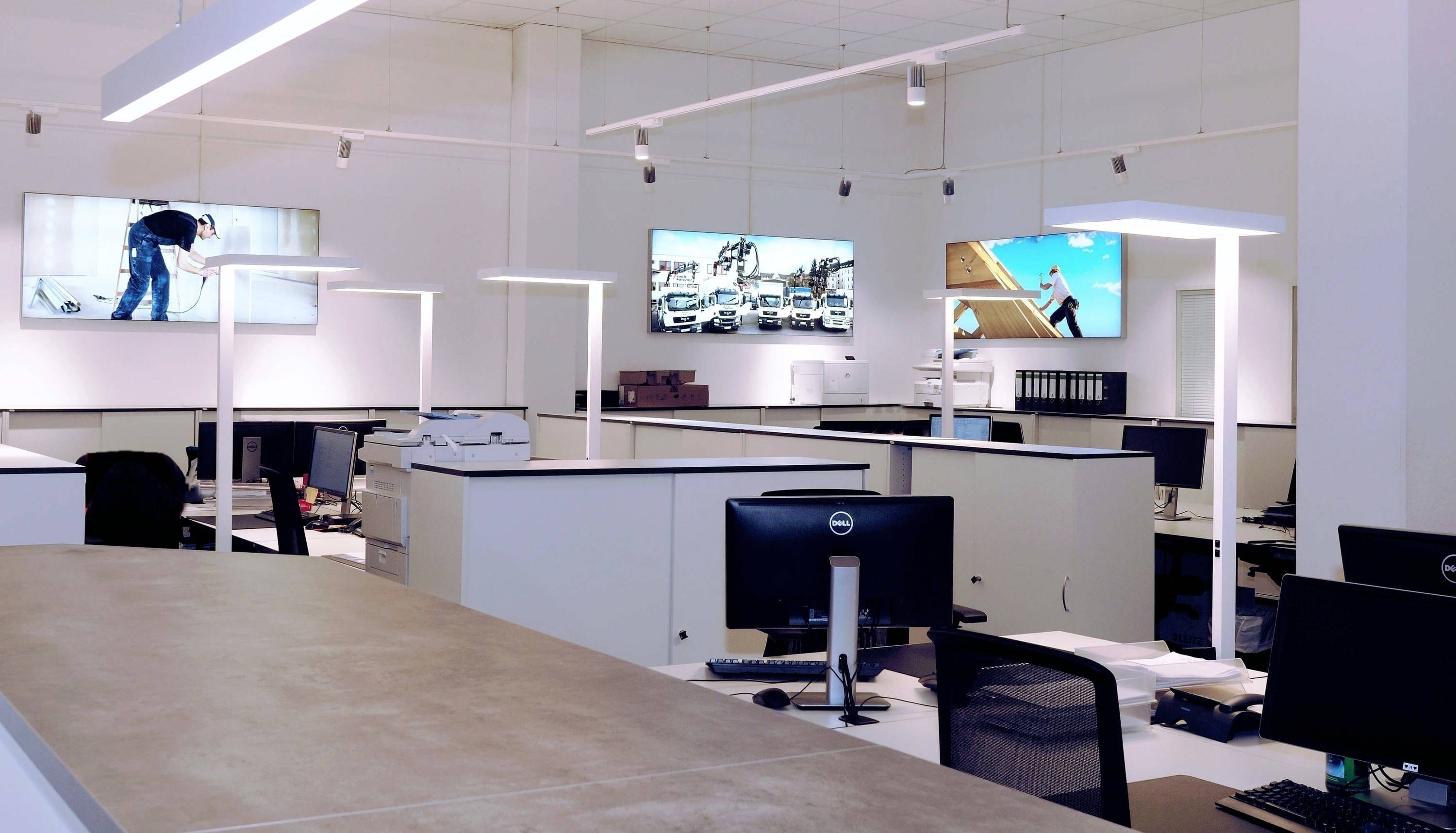 Moderne Bürofläche mit mehreren Schreibtischen, Computern und an der Wand montierten Bildschirmen, die in heller Umgebung Fotos zeigen.