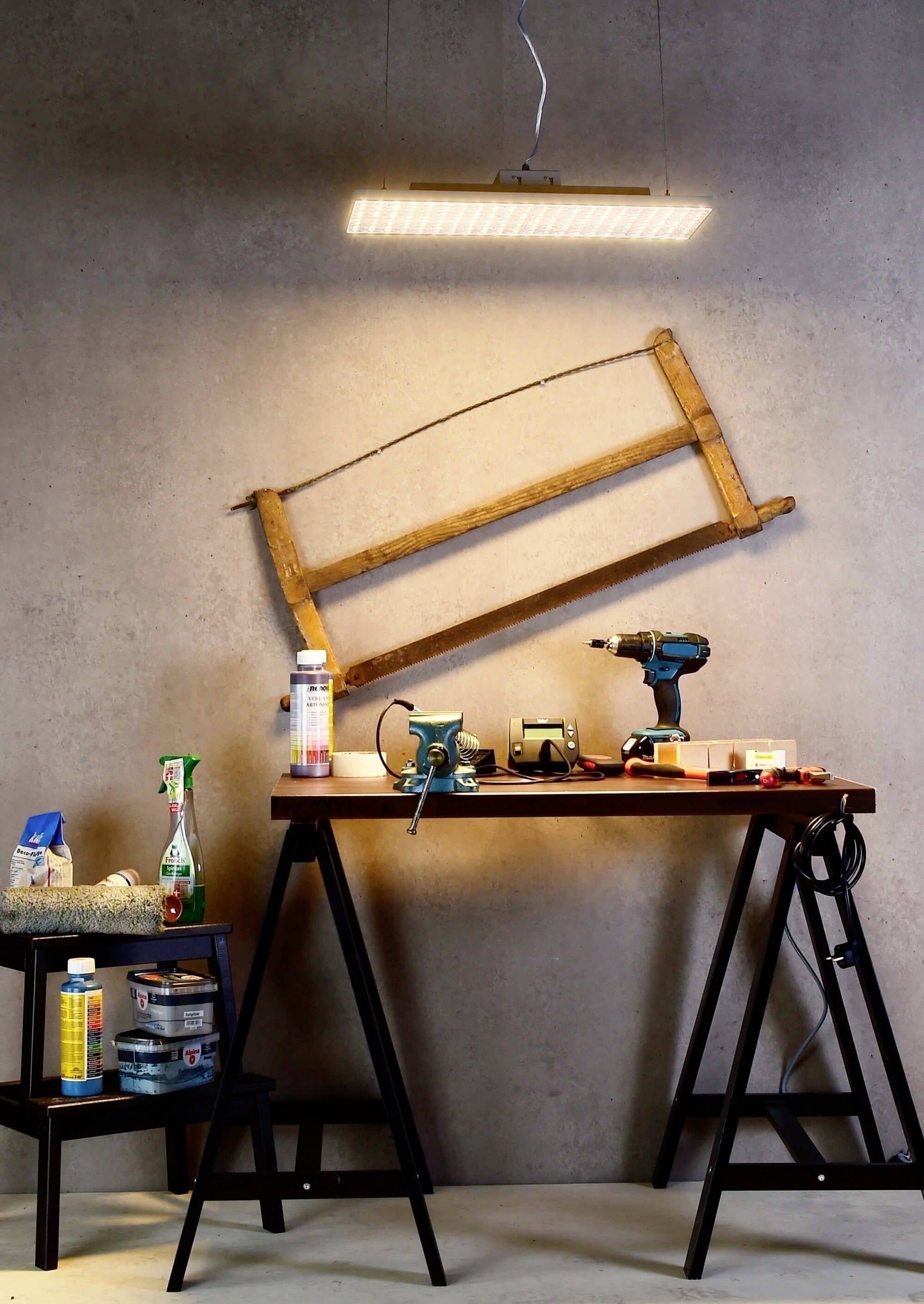 Arbeitsbereich mit Tisch; darauf liegen Werkzeug und Materialien wie Bohrmaschine und Schraubenschlüssel. An der Wand hängt ein alter Holzrahmen.