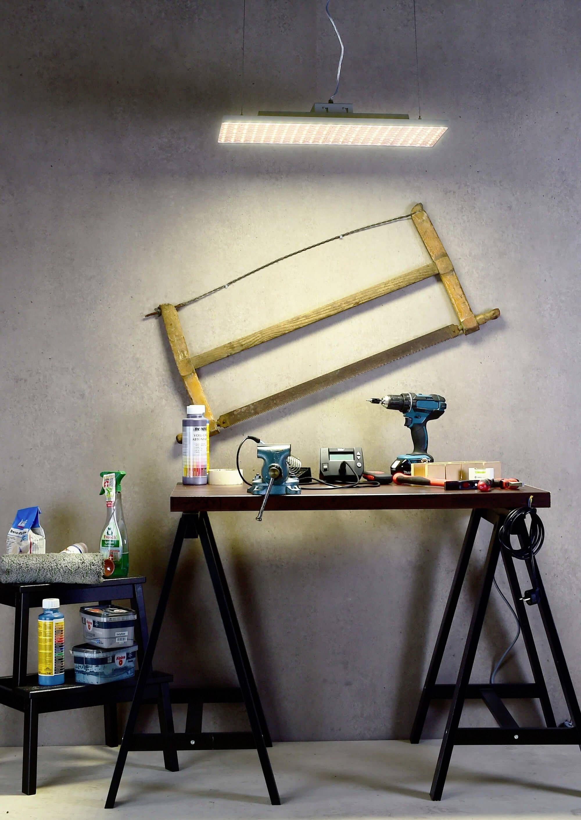 Eine Werkbank mit Werkzeugen und Materialien, darunter Bohrmaschine, Klebeband, Spraydosen und eine Lampe über einem hölzernen Rahmen an der Wand.