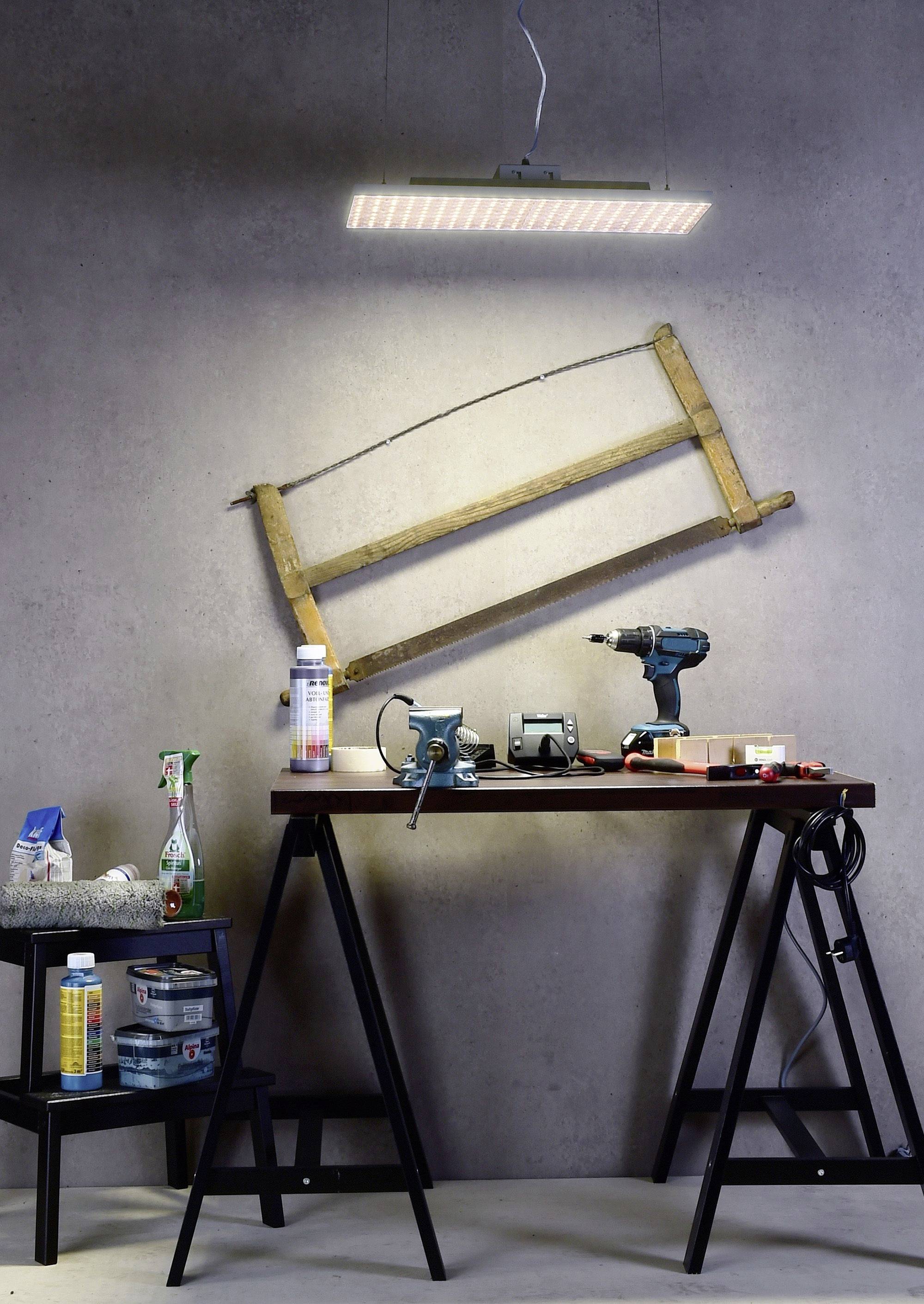 Werkbank mit Werkzeugen und Zubehör wie Bohrmaschine, Schleifgerät und Spraydosen unter einer Lampe, vor einer grauen Wand.