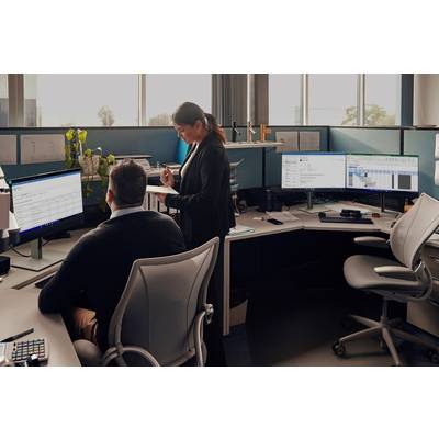 Eine Frau zeigt einem Mann an einem Schreibtisch etwas auf einem Bildschirm in einem modernen Büro mit mehreren Monitoren.