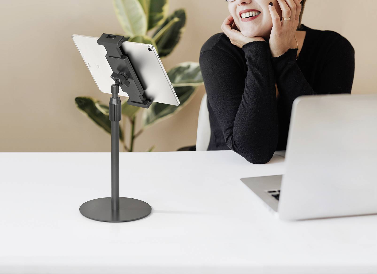 Eine Person sitzt an einem Tisch und lächelt, während sie auf einen Laptop und ein Tablet schaut, das auf einem Ständer befestigt ist. Im Hintergrund ist eine Pflanze.