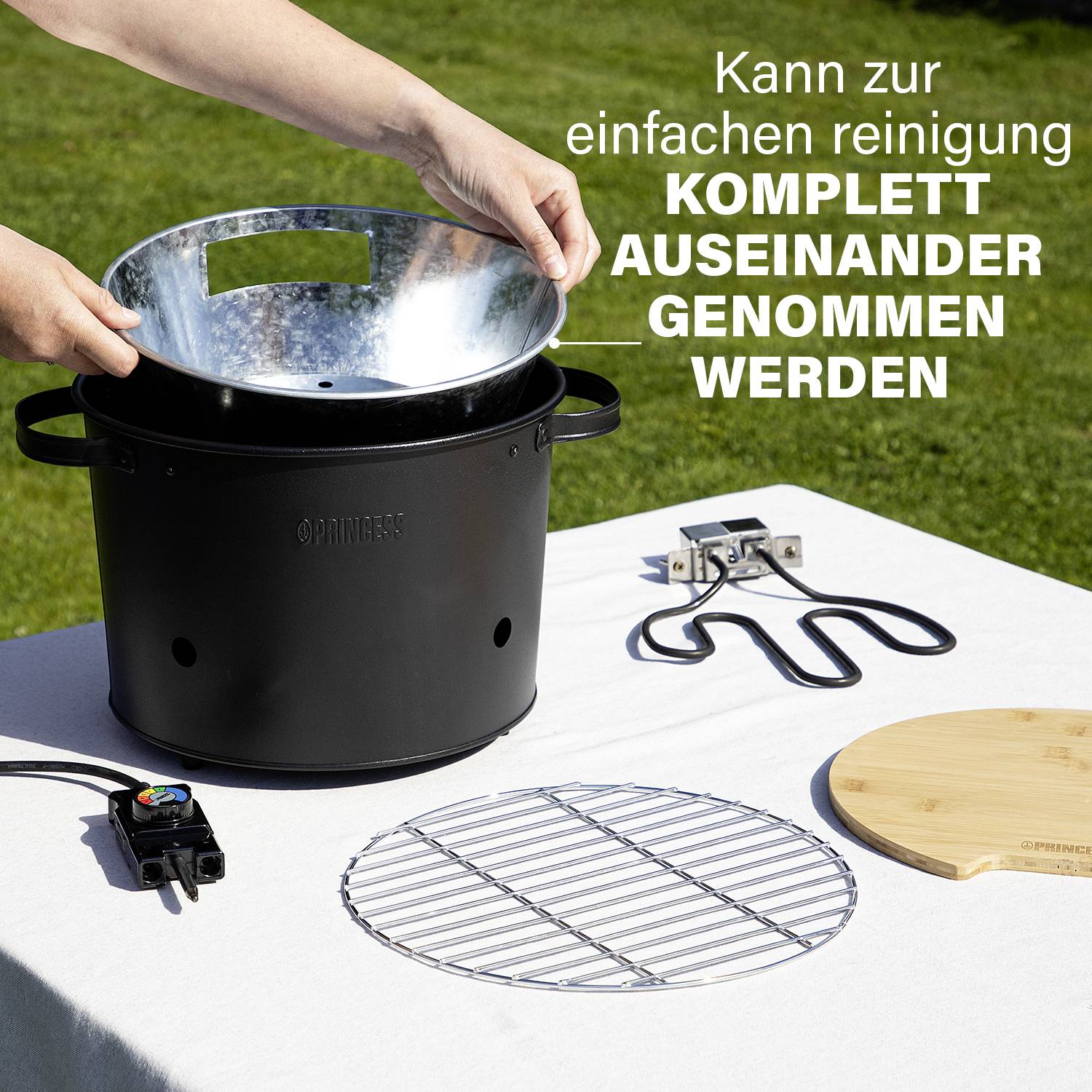 Eine Person zerlegt einen tragbaren Grill im Garten. Aufschrift: 'Einfach zu reinigen, KOMPLETT AUSEINANDER GENOMMEN WERDEN'. Teile liegen daneben.