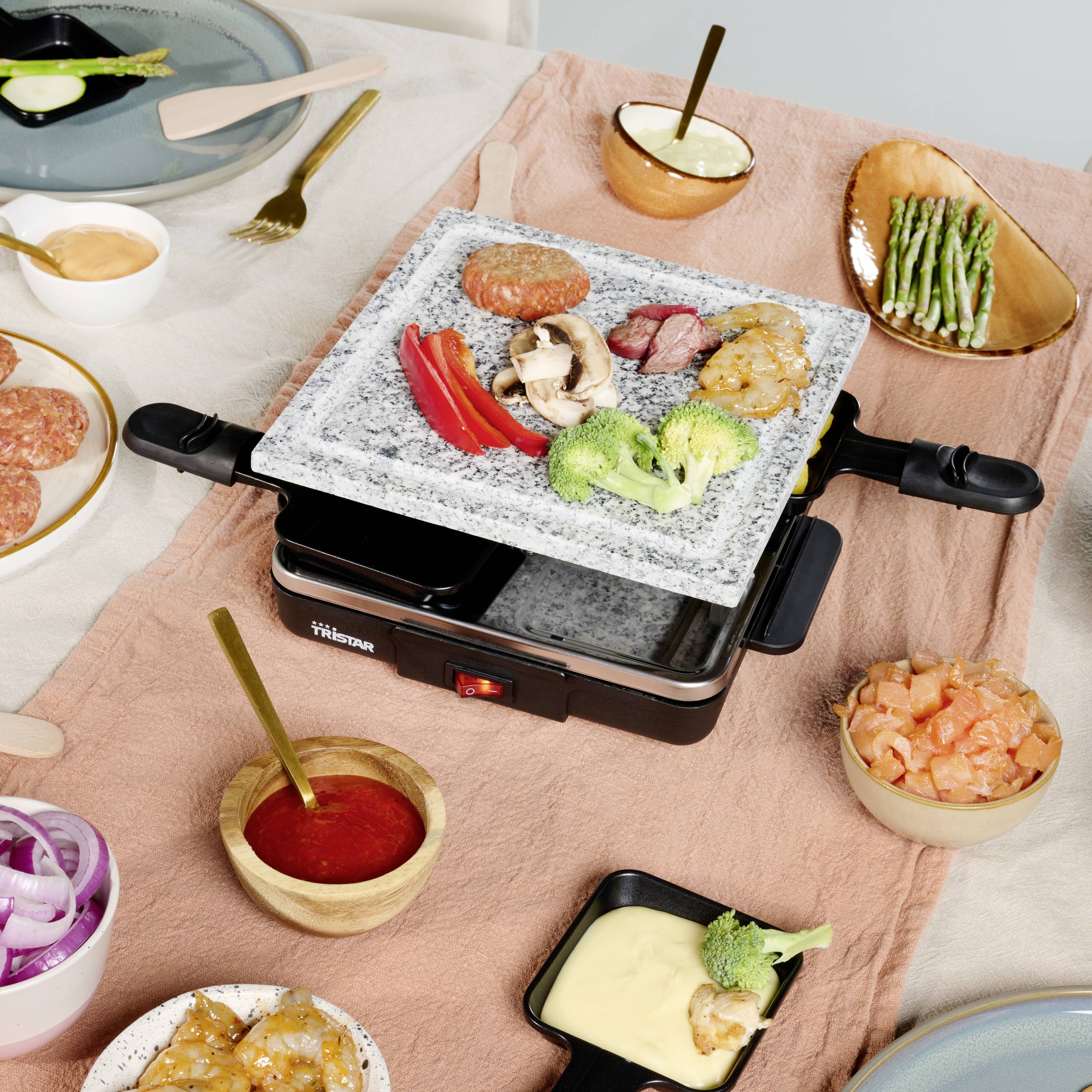 Ein Tisch mit Raclette-Grill und verschiedenen Zutaten, darunter Gemüse, Fleisch und Saucen. Bereit zur Zubereitung von Mahlzeiten.
