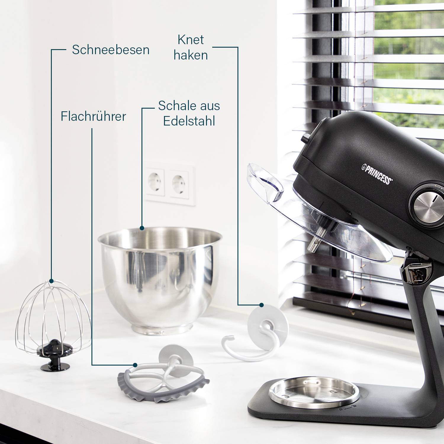 Küchenmaschine mit Zubehör: Schneebesen, Flachrührer, Knethaken und Edelstahlschale auf einem Küchentresen vor einer Jalousie.