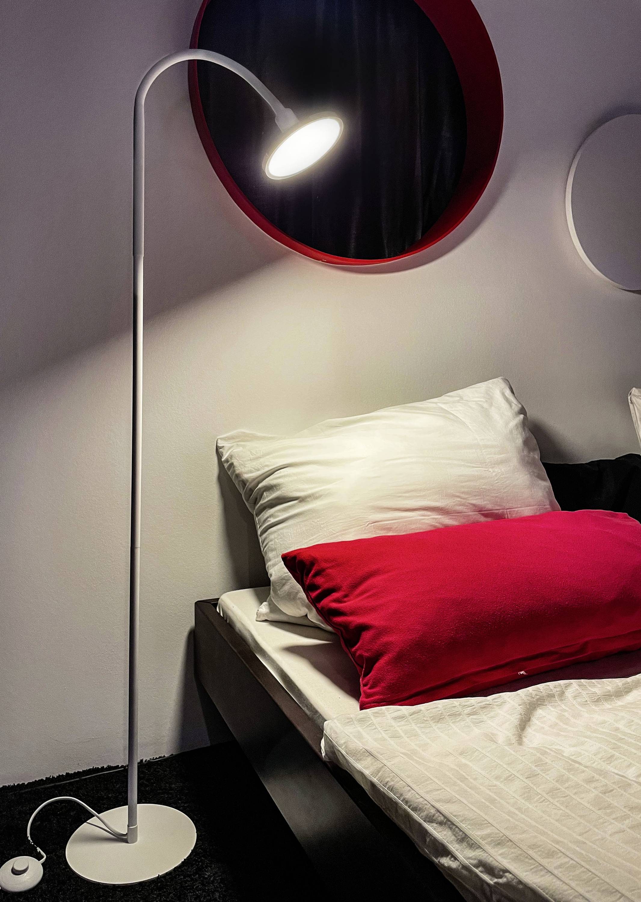 Stehlampe leuchtet über einem Bett mit weißen und roten Kissen. Schwarzer Teppich auf dem Boden. Runder Spiegel an der Wand.