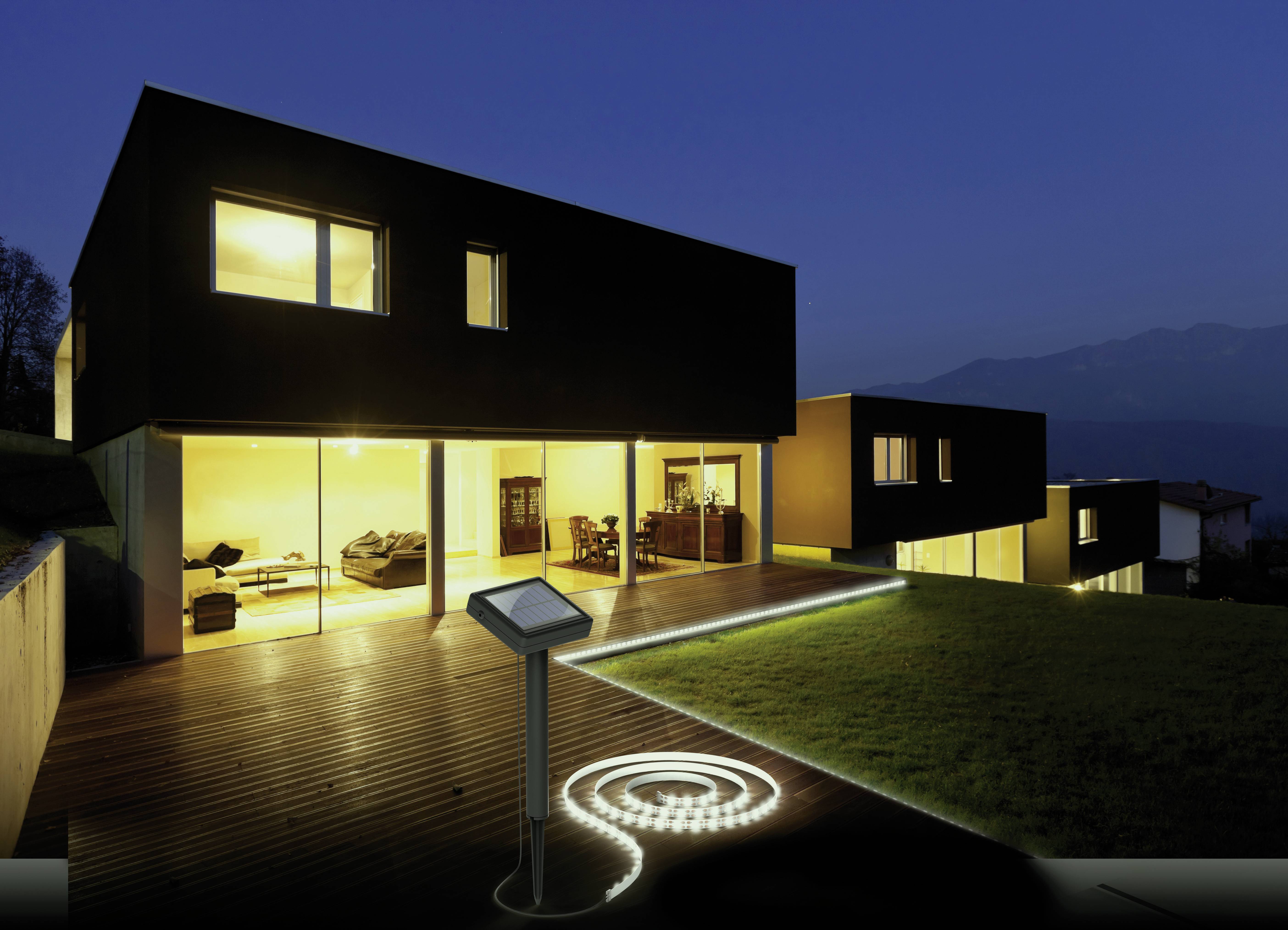 Ein modernes, quadratisches Haus mit großen, erleuchteten Fenstern bei Nacht, umgeben von einem gepflegten Garten mit beleuchtetem Pfad.