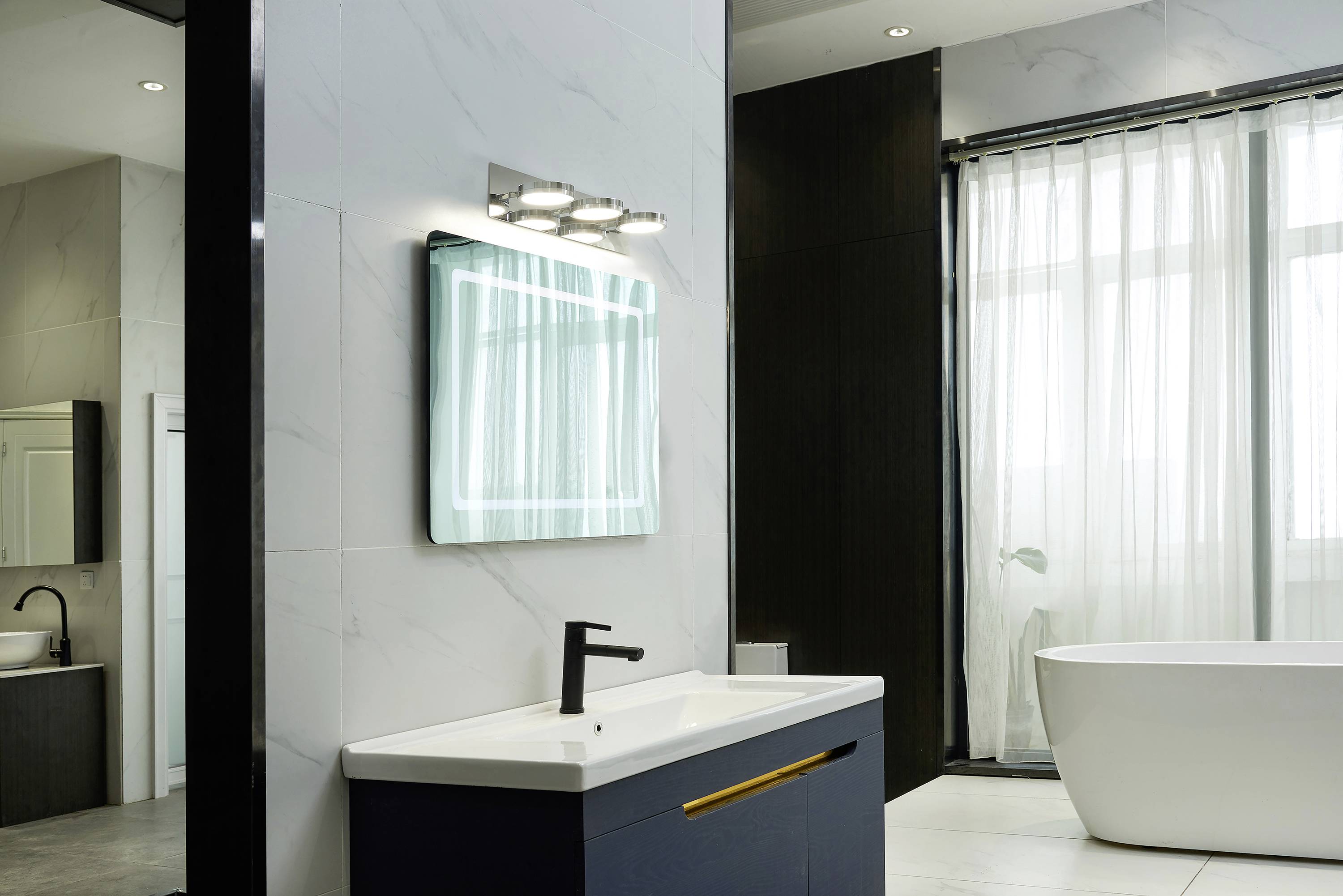 Modernes Badezimmer mit wandmontiertem Waschbecken, beleuchtetem Spiegel und freistehender Badewanne vor einem großen Fenster.