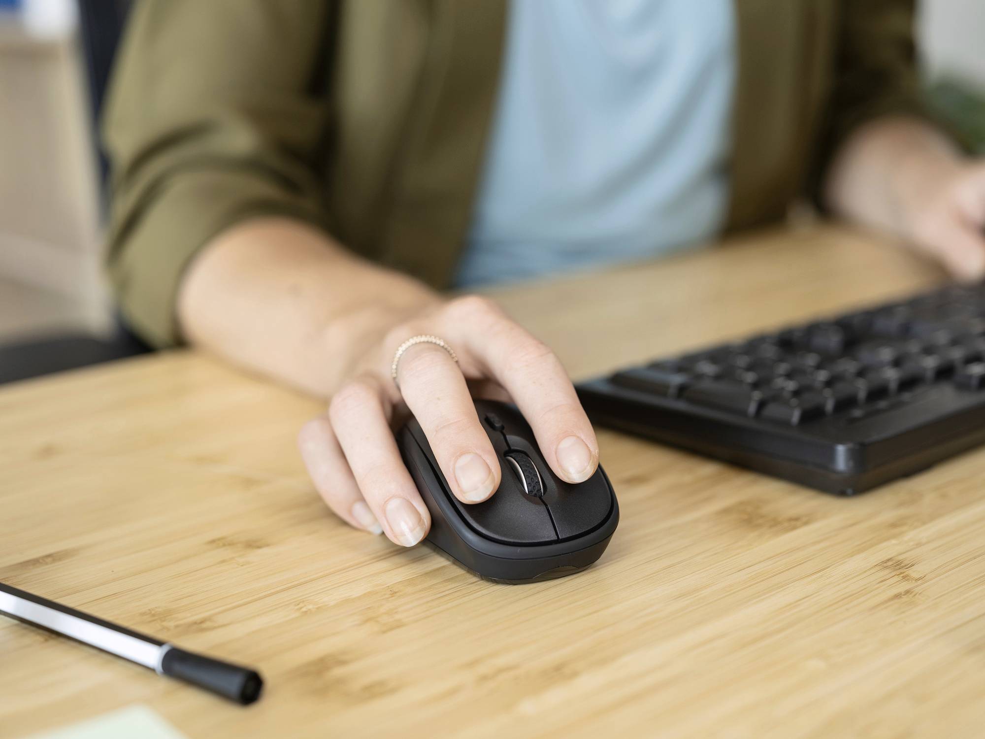 Eine Person bedient eine Computermaus auf einem Holztisch, neben einer Tastatur und einem Stift. Szene zeigt Arbeit am Schreibtisch.