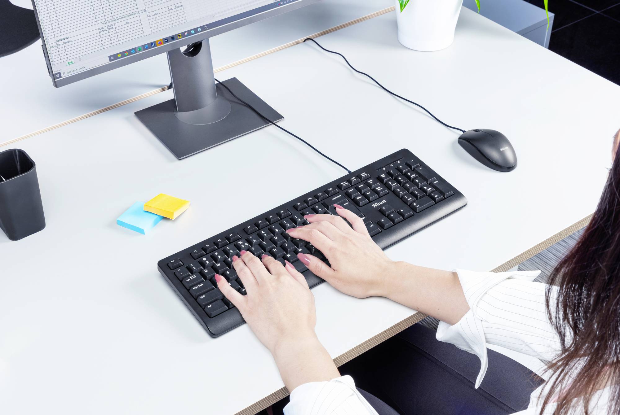 Eine Person tippt auf einer Tastatur an einem Schreibtisch mit einem Monitor. Auf dem Tisch liegen Klebezettel und ein Stift.