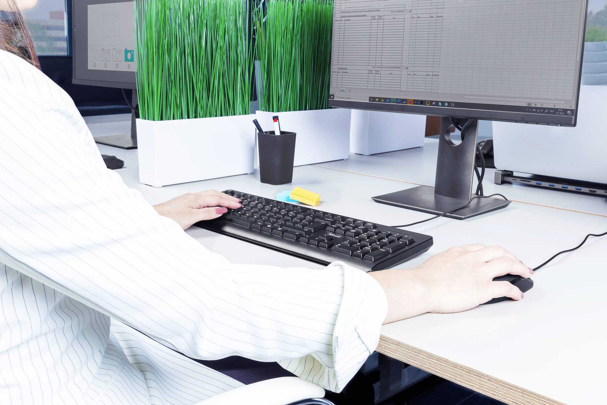 Eine Person arbeitet an einem Computer mit zwei Monitoren auf einem Schreibtisch mit Pflanzen und Bürozubehör.