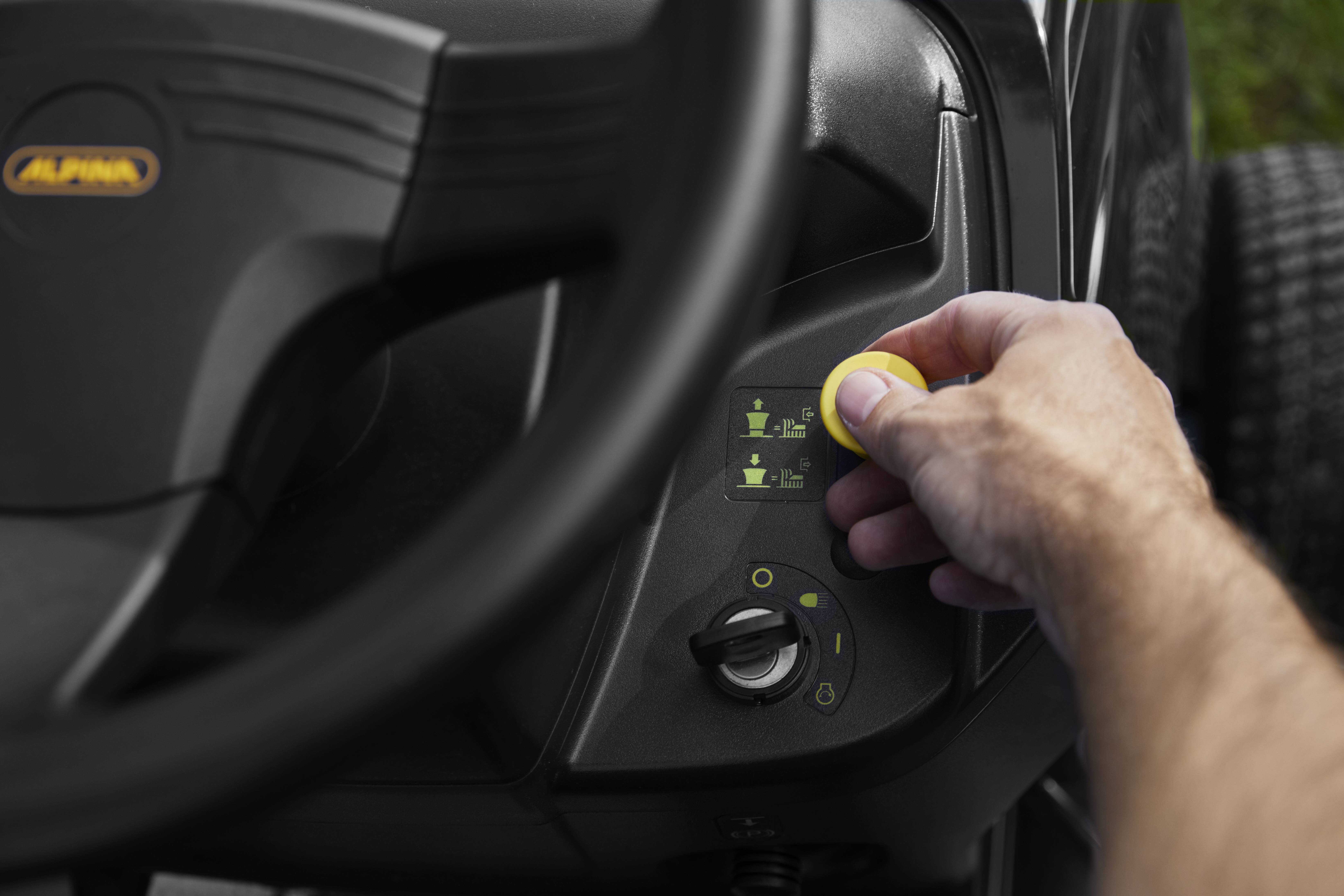 Eine Person bedient die Steuerkonsole eines Rasentraktors, indem sie einen gelben Knopf dreht, um die Schnitthöhe einzustellen.