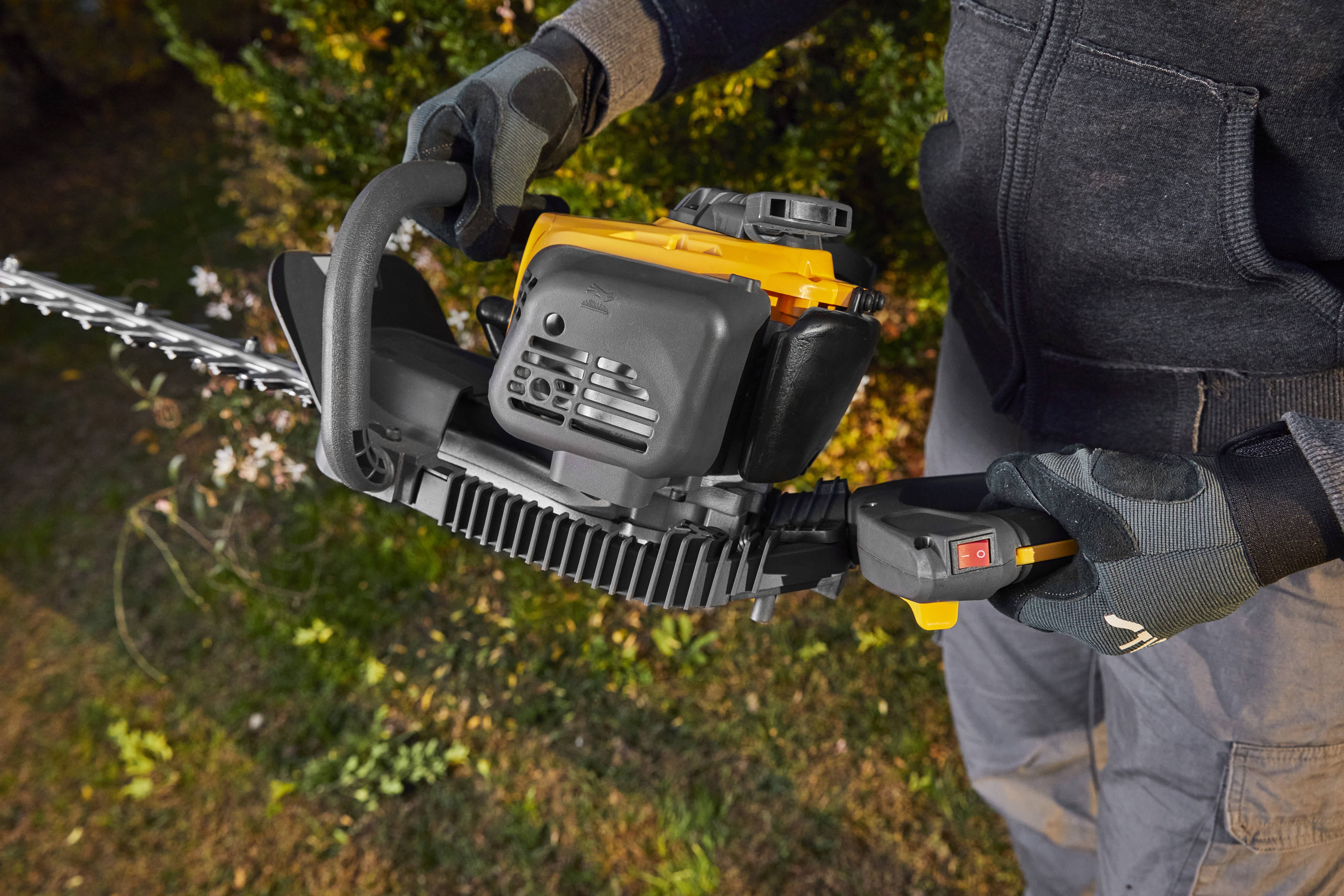 Person mit Handschuhen schneidet mit einer gelben Heckenschere eine Hecke.