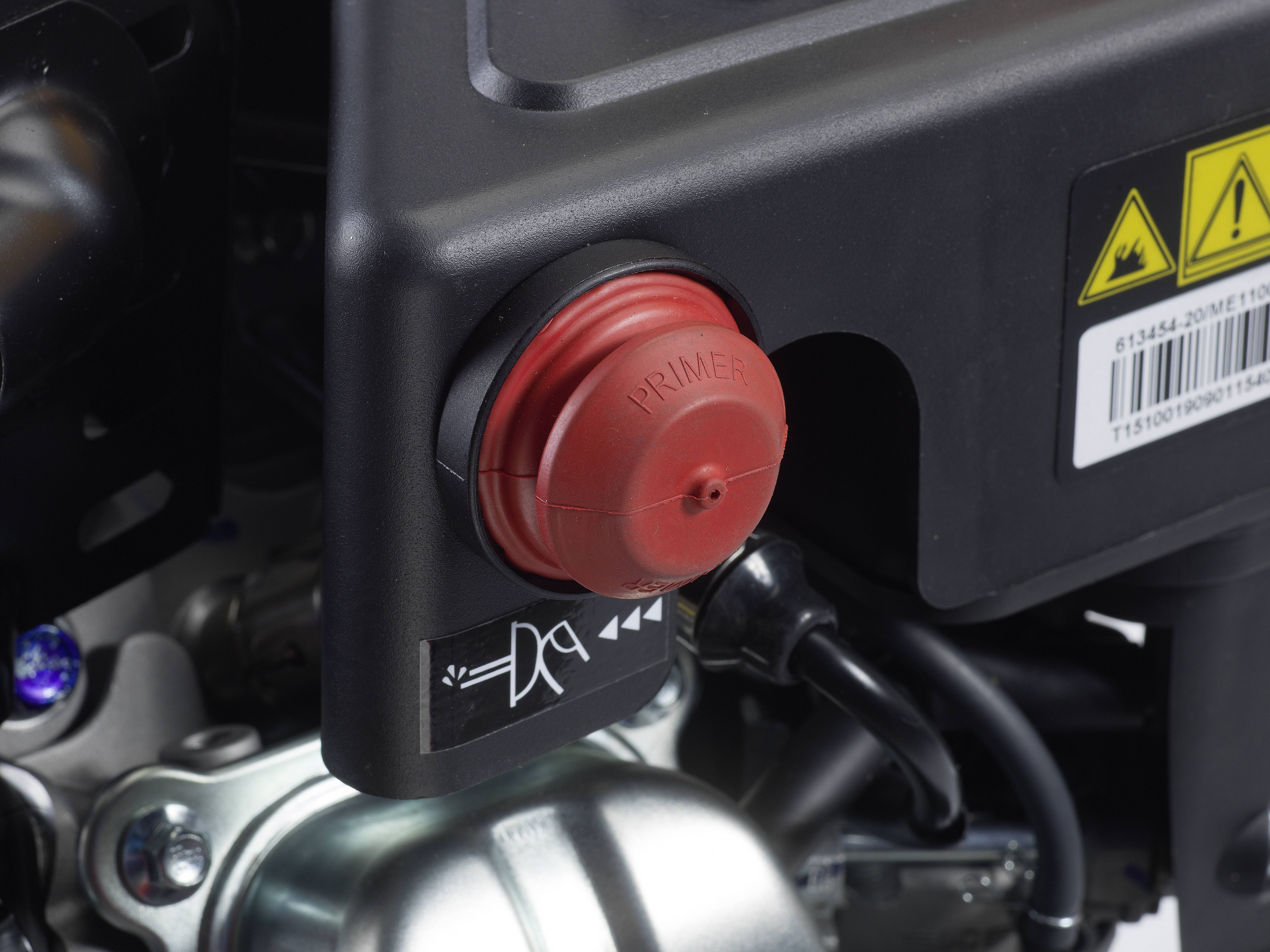 Roter Knopf mit der Aufschrift 'Primer' auf einer Maschine. Darunter sind Symbole für Bedienung. Im Hintergrund Maschinenbauteile sichtbar.