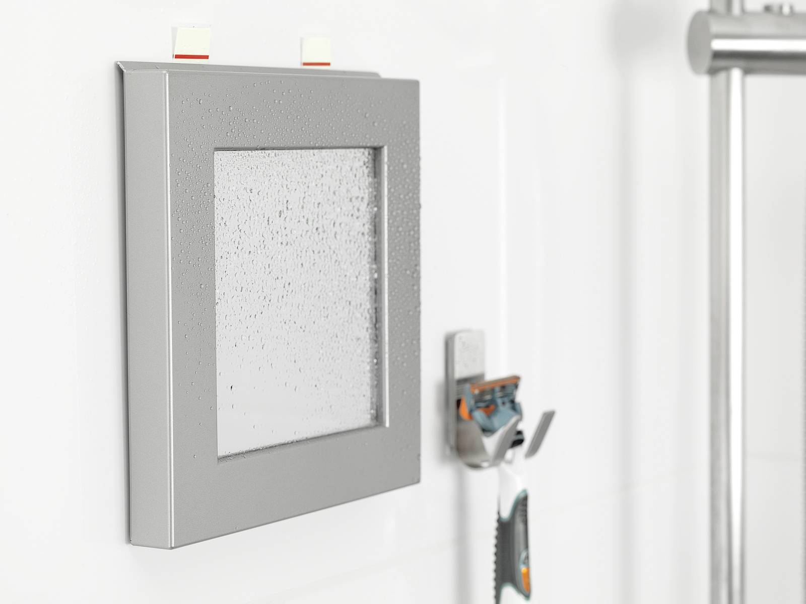 Spiegel mit grauem Rahmen und Wassertröpfchen an der Wand eines Badezimmers, daneben ein Rasierer auf einer Halterung.