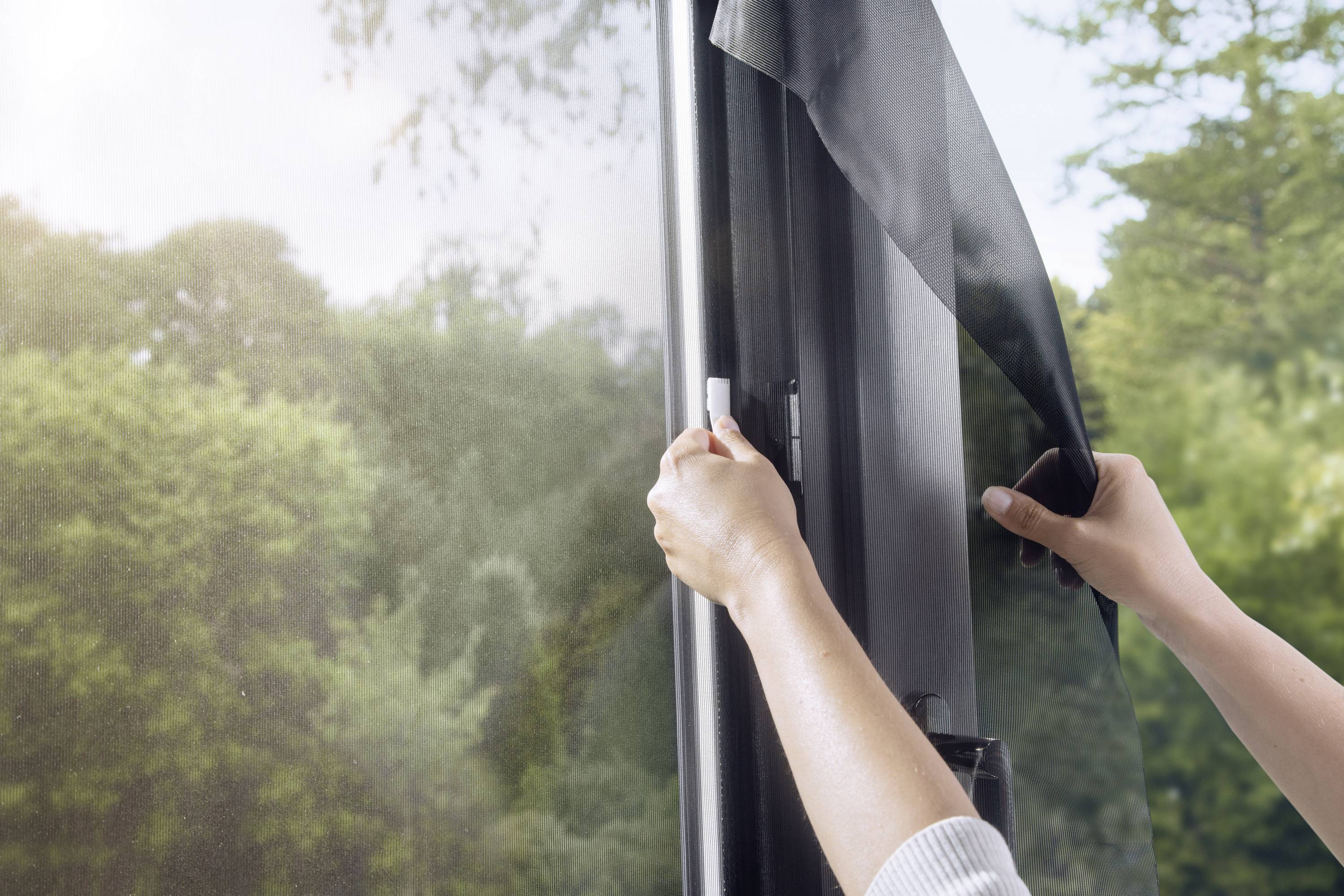Eine Person bringt eine Sonnenschutzfolie an einem Fenster an, um den Lichteinfall zu reduzieren. Im Hintergrund sind Bäume sichtbar.