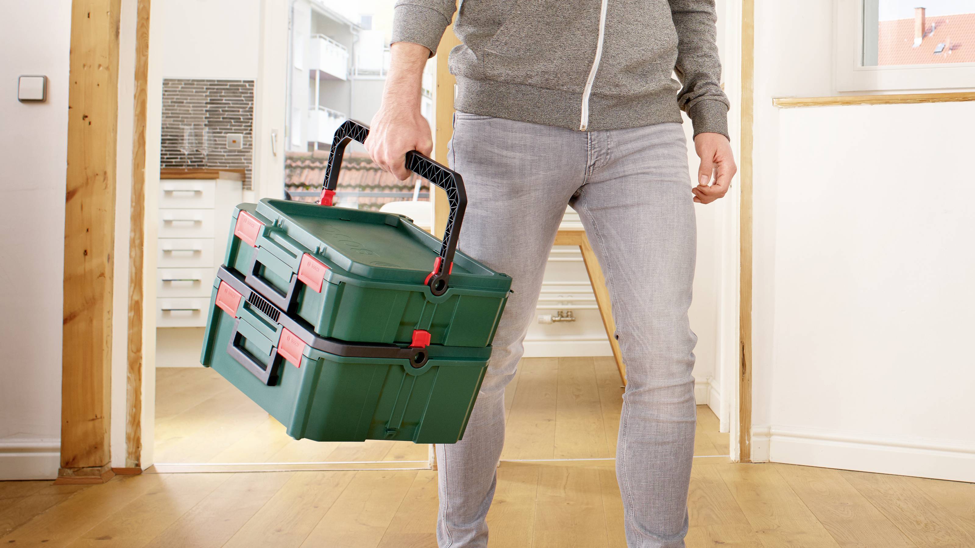 Eine Person trägt einen grünen Werkzeugkasten mit zwei Griffen in einem hellen Raum. Im Hintergrund sind Möbel und eine Tür zu sehen.