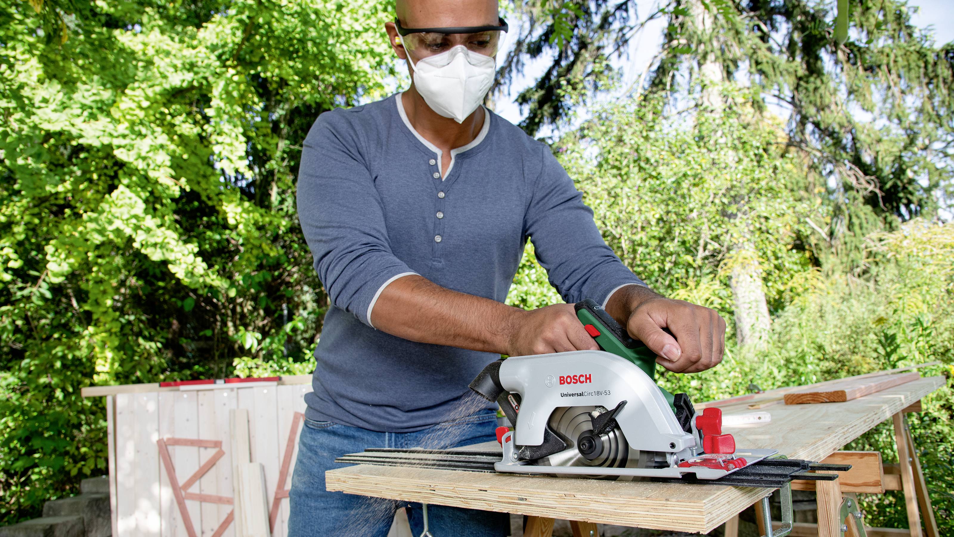Eine Person schneidet mit einer Bosch-Kreissäge Holz im Freien, trägt Schutzbrille und Maske. Hintergrund zeigt grüne Bäume.