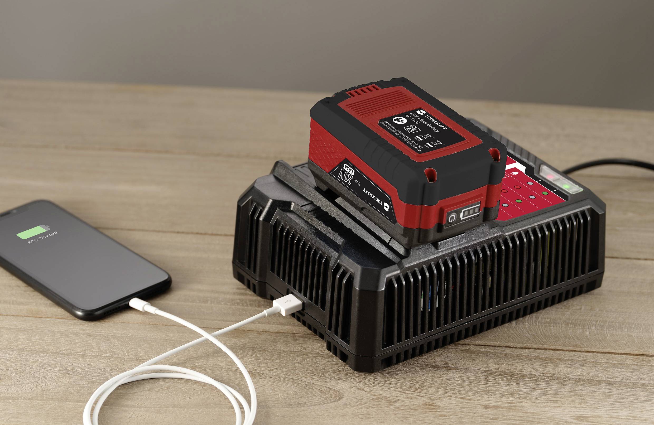 Ein Akkuladegerät lädt einen roten Akku. Ein Smartphone mit Ladeanzeige ist über ein Kabel mit dem Ladegerät verbunden.