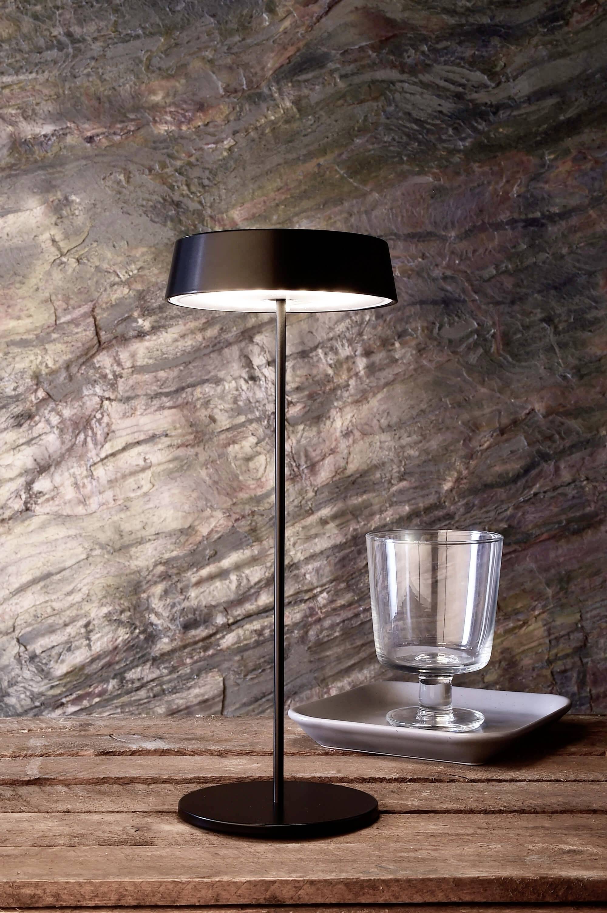 Eine elegante schwarze Tischlampe auf einem Holztisch, neben einem leeren Glas auf einem quadratischen Teller, vor steiniger Wand.