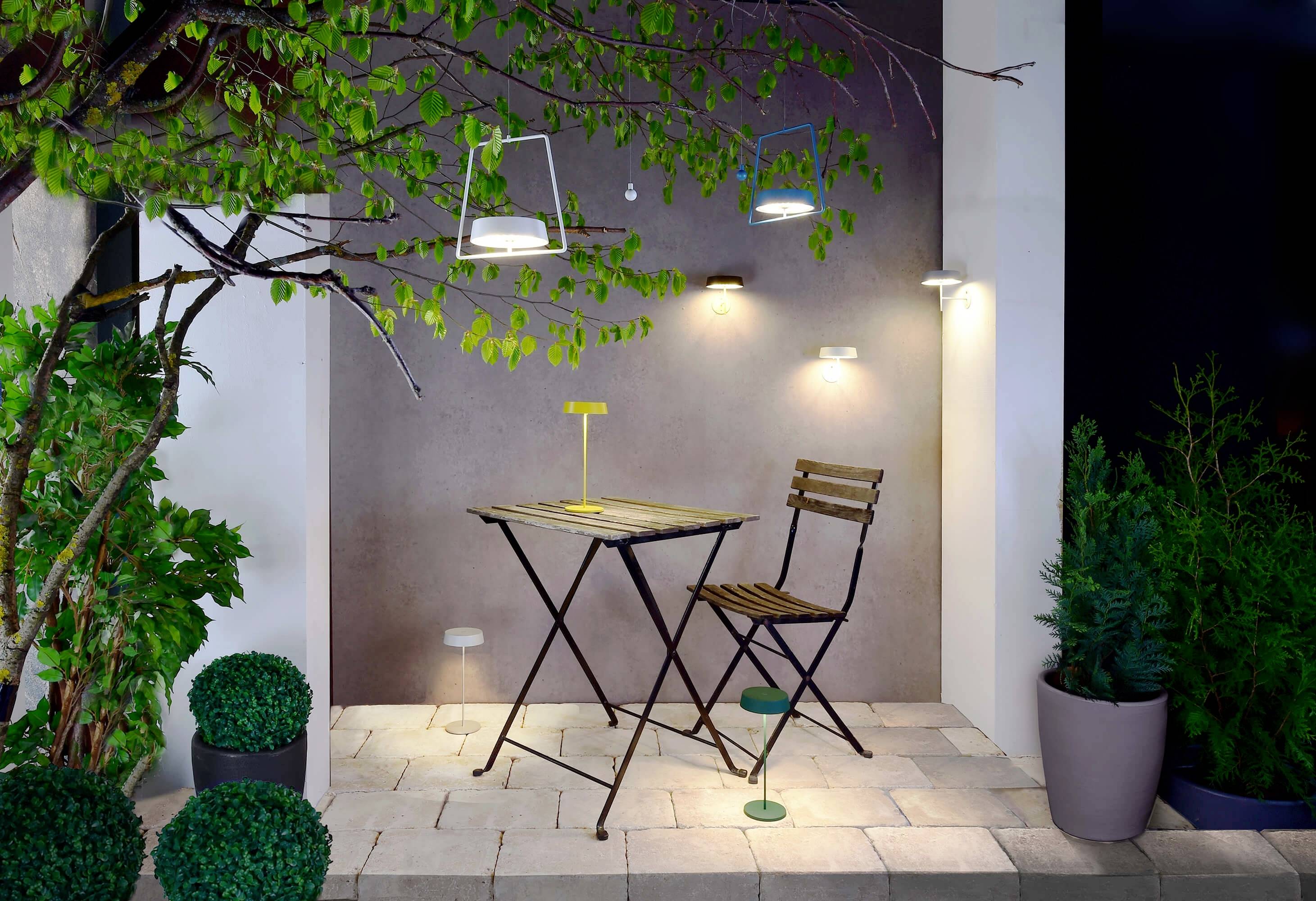 Ein kleiner Balkon mit Tisch und Stuhl, umgeben von Pflanzen. An der Wand hängen zwei Außenlampen, die den Bereich beleuchten.