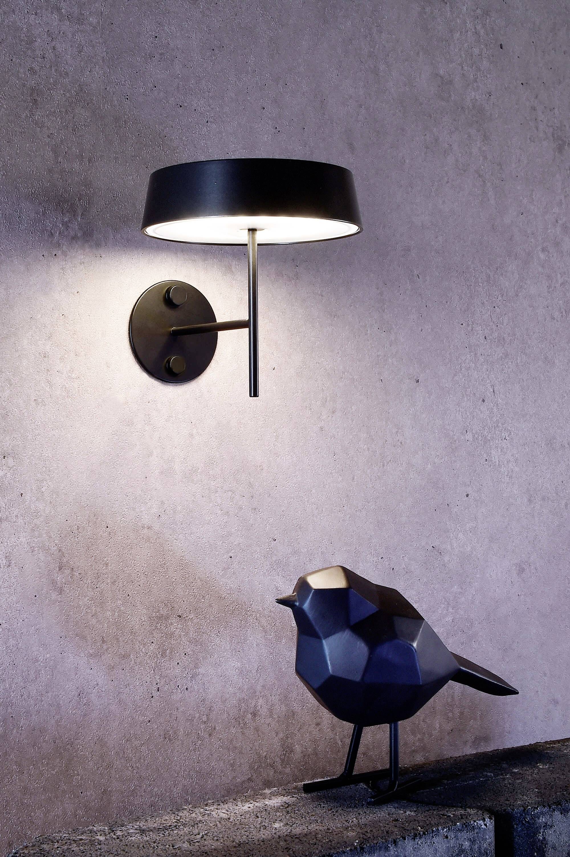 Eine moderne, schwarze Wandleuchte strahlt warmes Licht auf eine stilisierte, geometrische Vogelstatue auf einem Betonsockel.