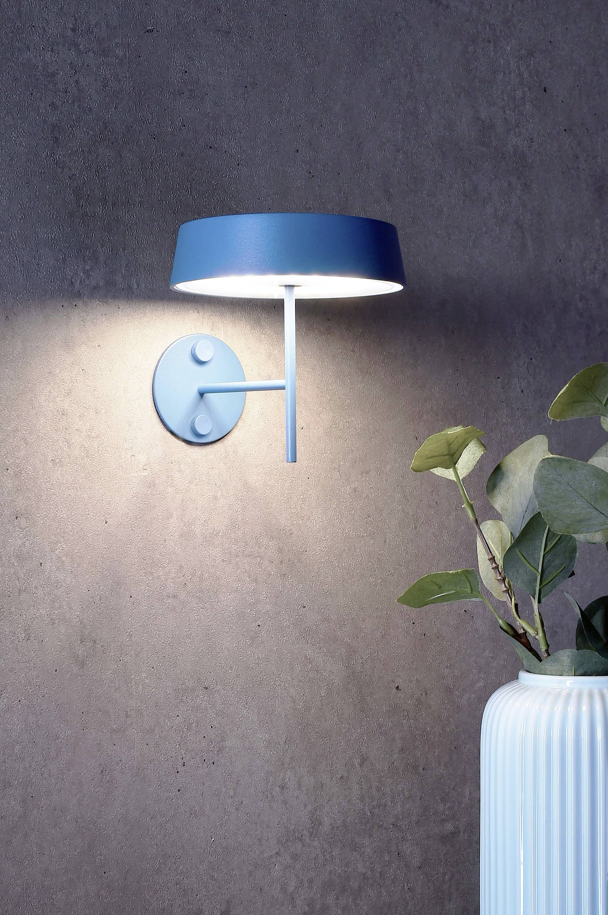 Blaue Wandleuchte, die nach unten auf eine strukturierte Wand leuchtet. Rechts darunter steht eine Vase mit grünen Blättern.