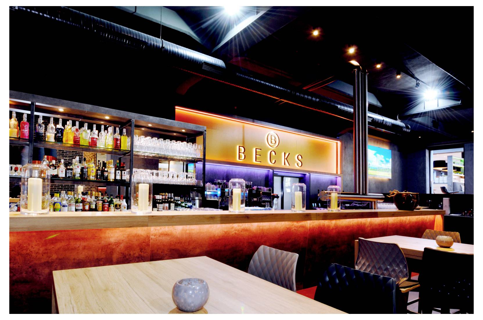 Moderne Bar-Innenausstattung mit leuchtendem „BECKS“-Schild, hinterleuchteten Spirituosenregalen, Holztischen und leeren Sitzplätzen unter industriellen Deckenleuchten.