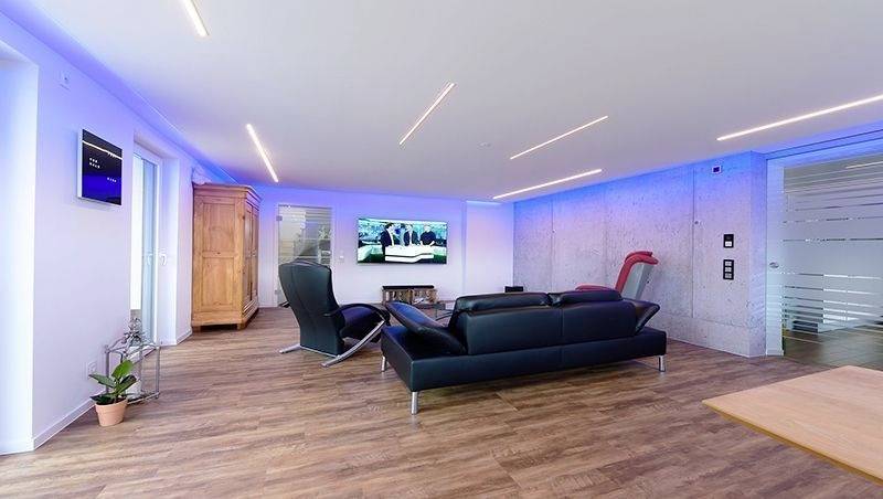 Ein modernes Wohnzimmer mit Holzfußboden, L-förmigem schwarzem Sofa, Sessel, Wand-TV und blauer Ambientebeleuchtung.