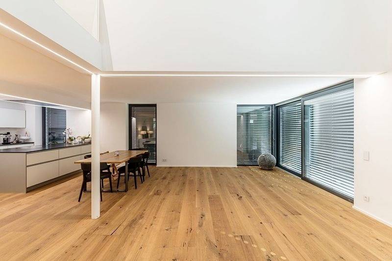 Großes Wohnzimmer mit Holzboden, modernem Essbereich, offener Küche links und großen Fenstern mit Jalousien rechts.