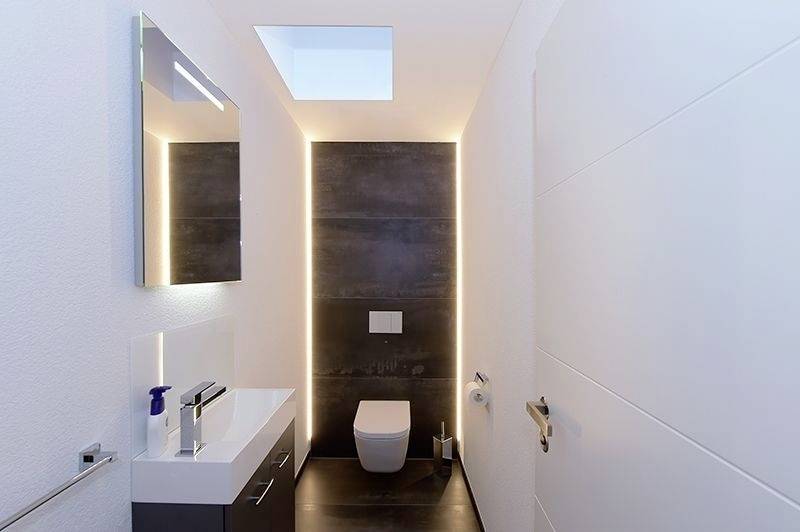 Modernes Badezimmer, schmaler Raum mit Toilette, Waschbecken und Spiegel. Beleuchtung betont die Wand hinter der Toilette.